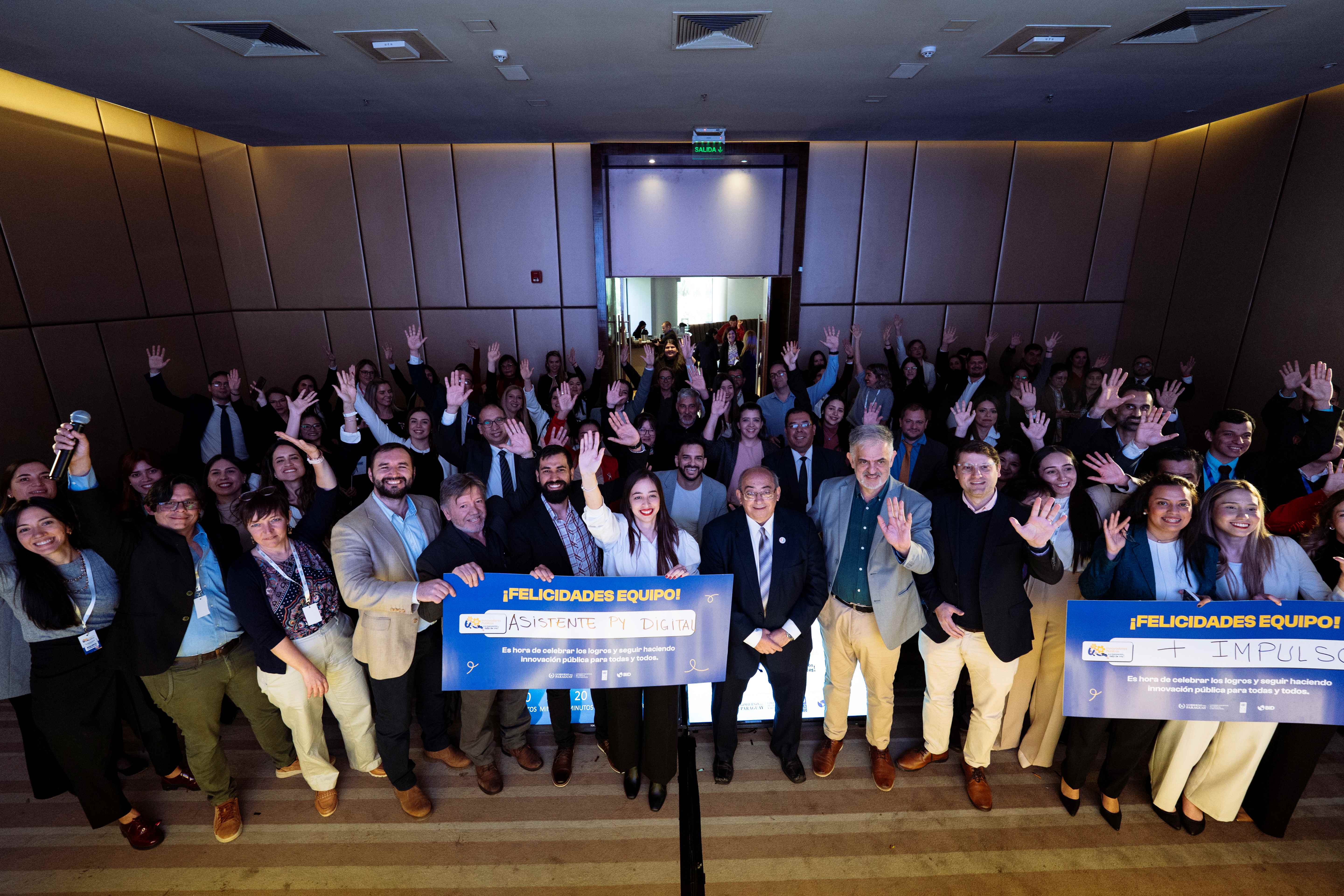 A large group of attendees raising their hands in a conference room, smiling.