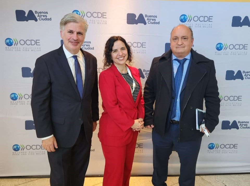 Tres personas posan para una foto en una conferencia, sonriendo delante de un banner con logos.