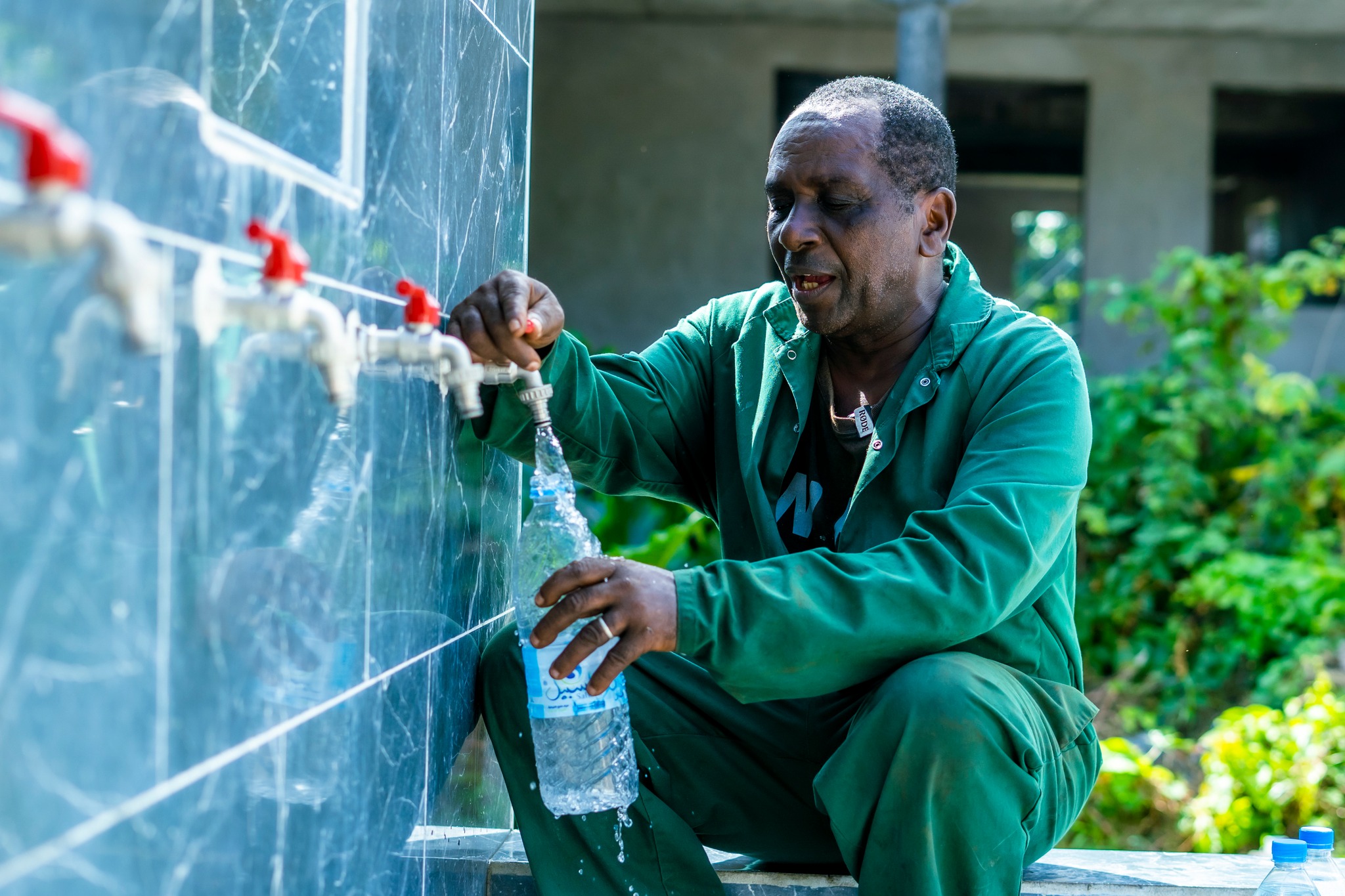 Man in green attire fills a bottle at a water tap, surrounded by greenery.