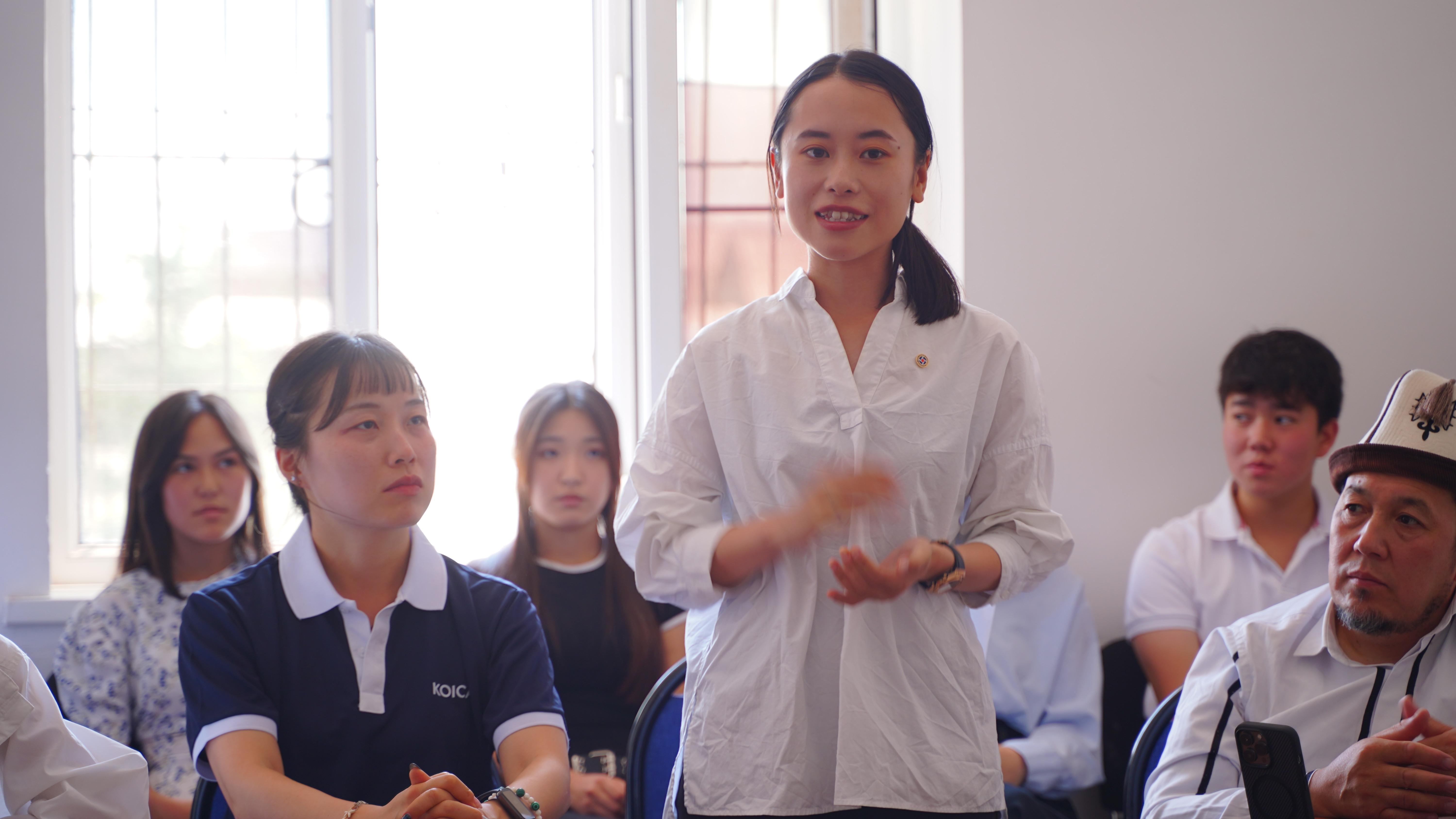 A woman in a white shirt gestures while speaking to a seated audience in a bright room.
