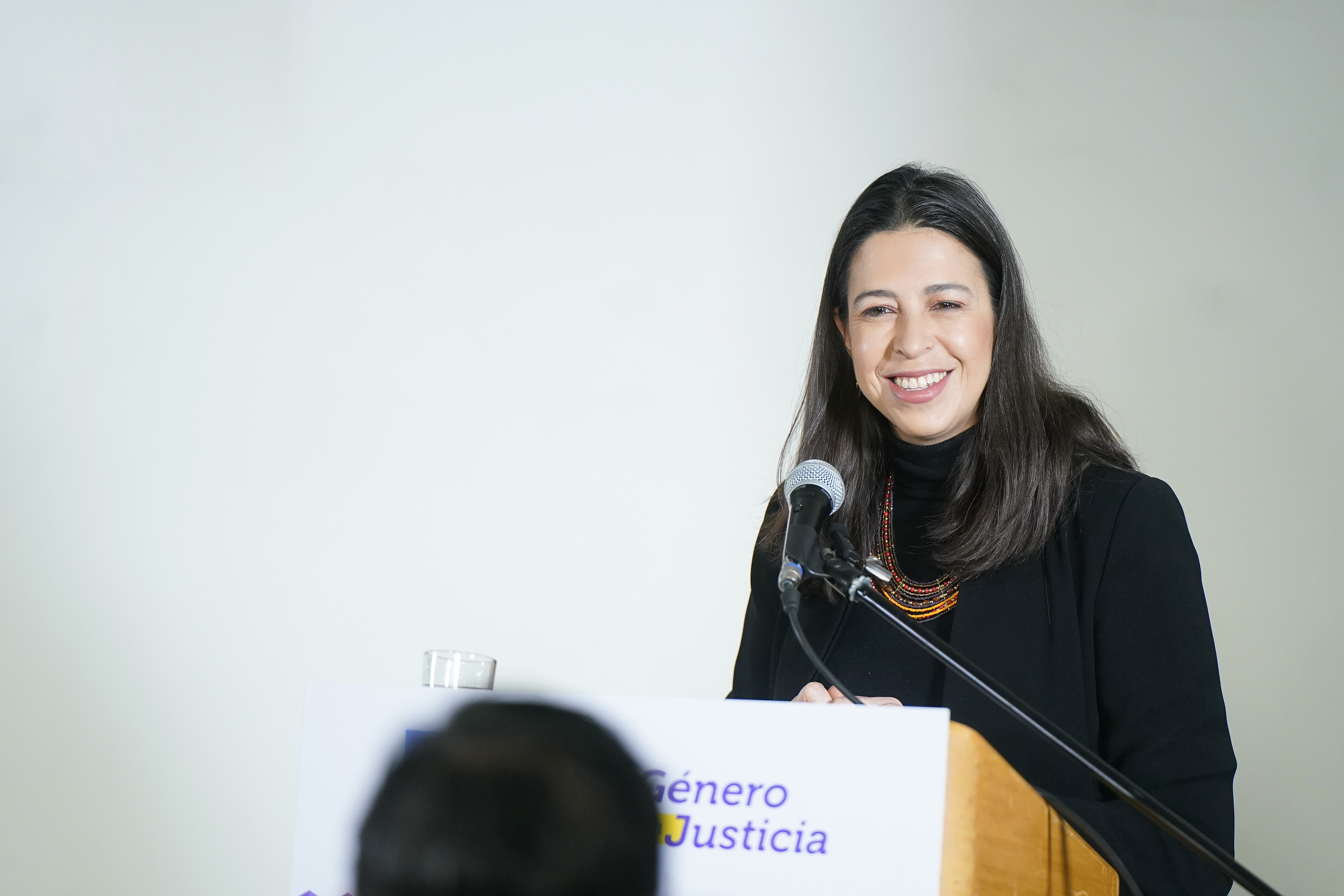 A woman smiles while speaking at a podium, with a microphone and a glass of water nearby.