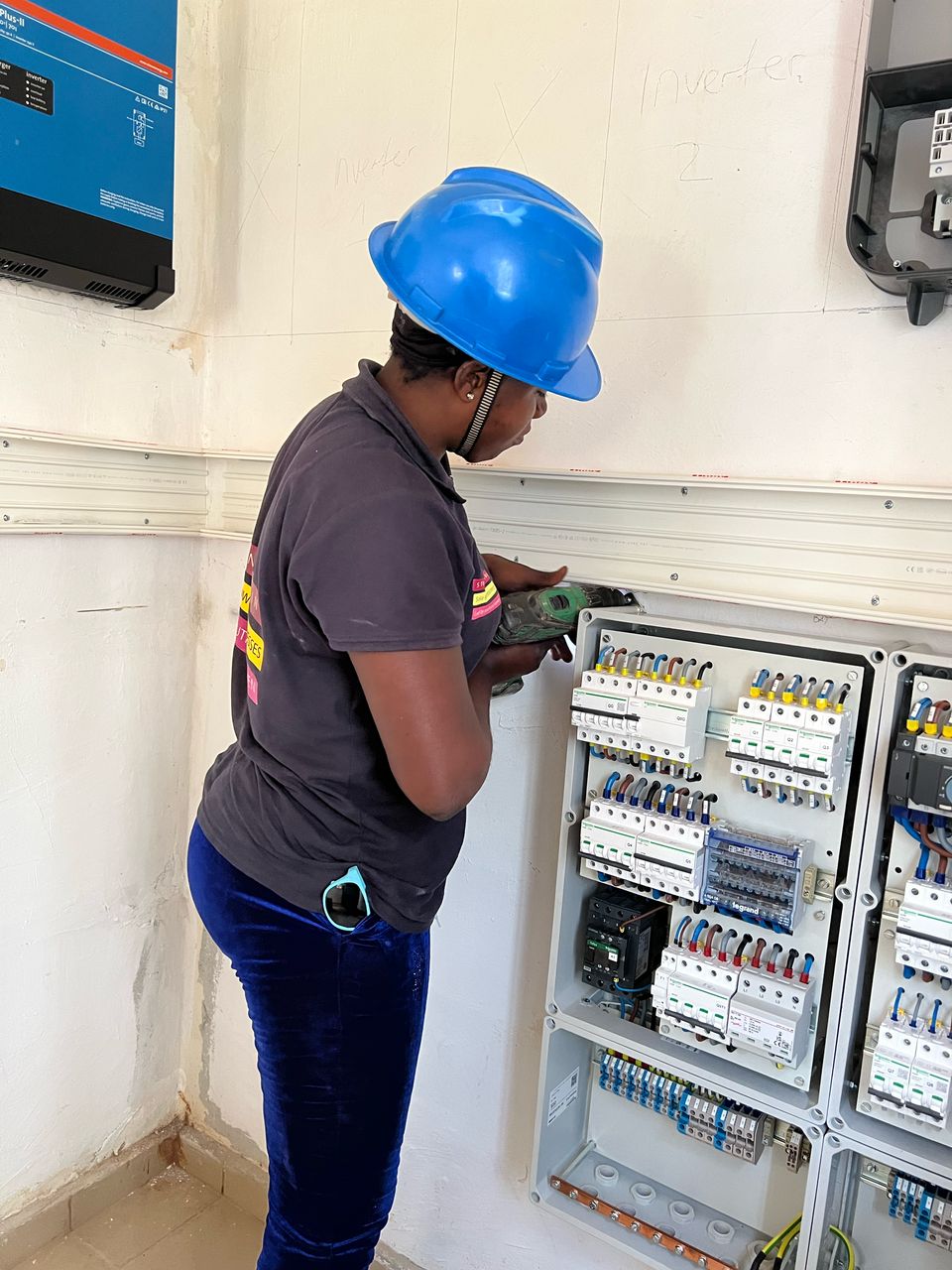 A woman in a blue hard hat works on an electrical panel, tools in hand.