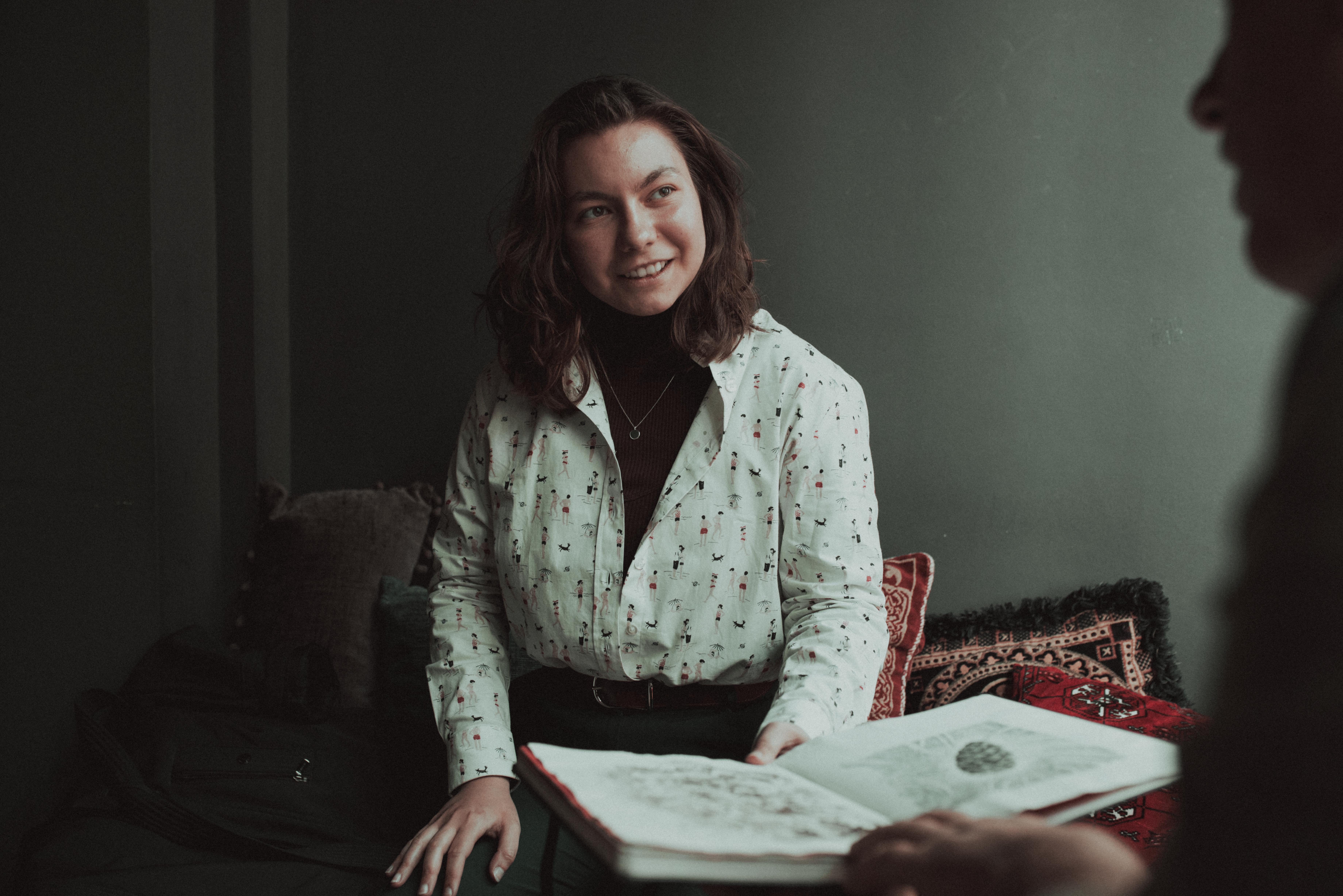A smiling woman in a floral shirt sits, engaging with a book in her hands.
