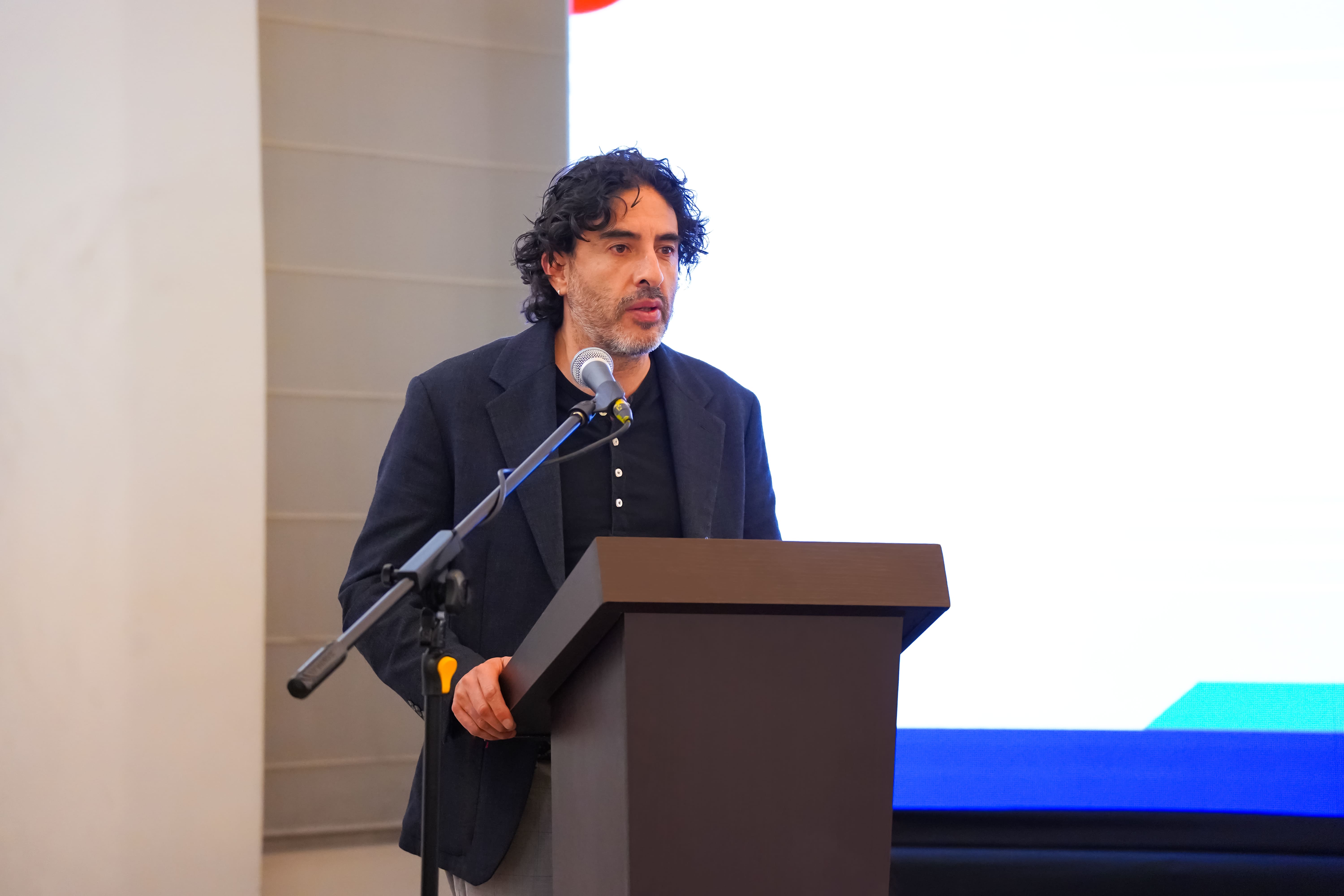A man with curly hair speaks at a podium with a microphone, screen behind him.