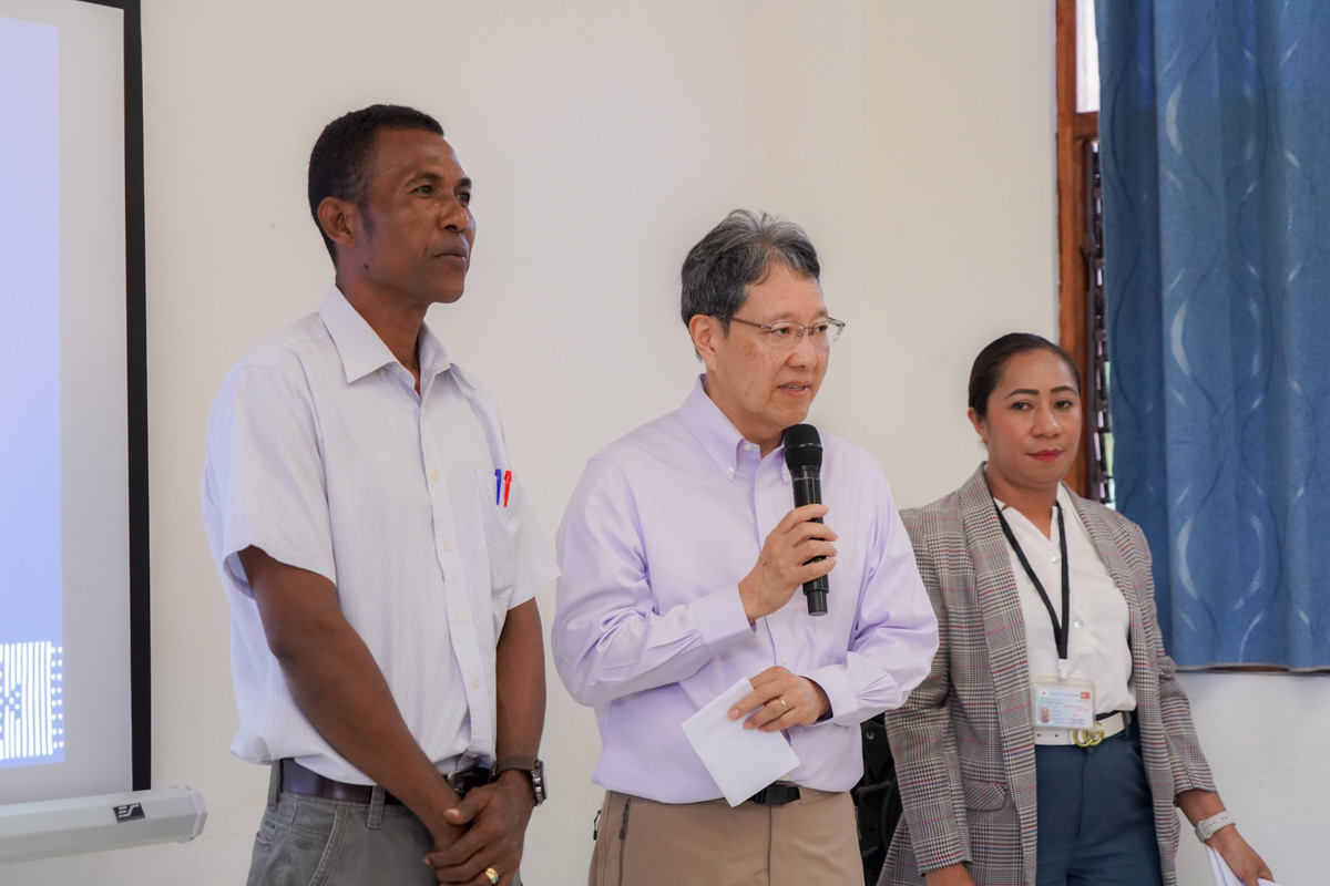 Three people stand in a room during a presentation, with one speaking into a microphone.