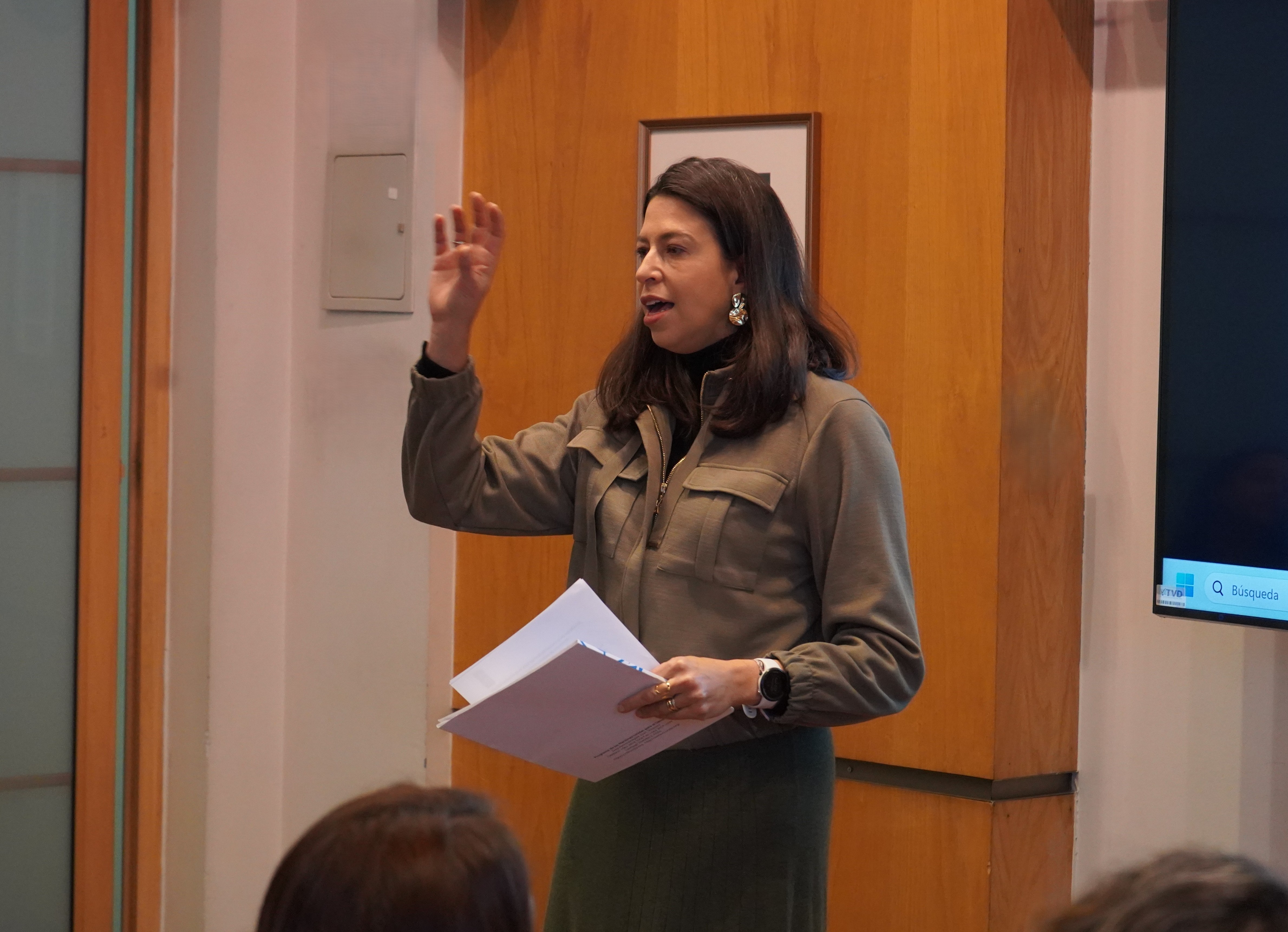A woman speaking passionately with one hand raised, holding papers, in a meeting room.
