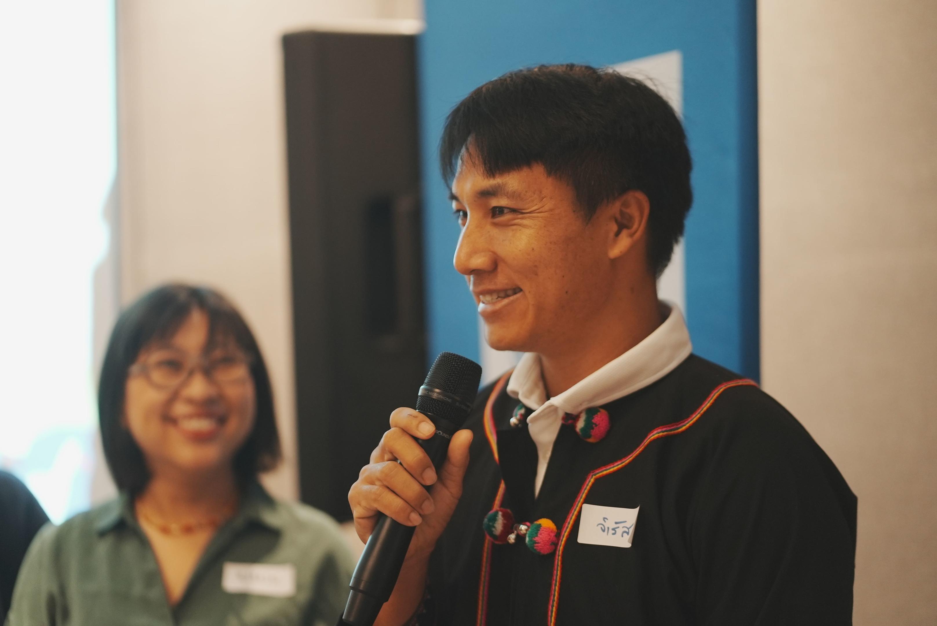 A man in traditional attire speaks into a microphone, smiling; a woman stands beside him.