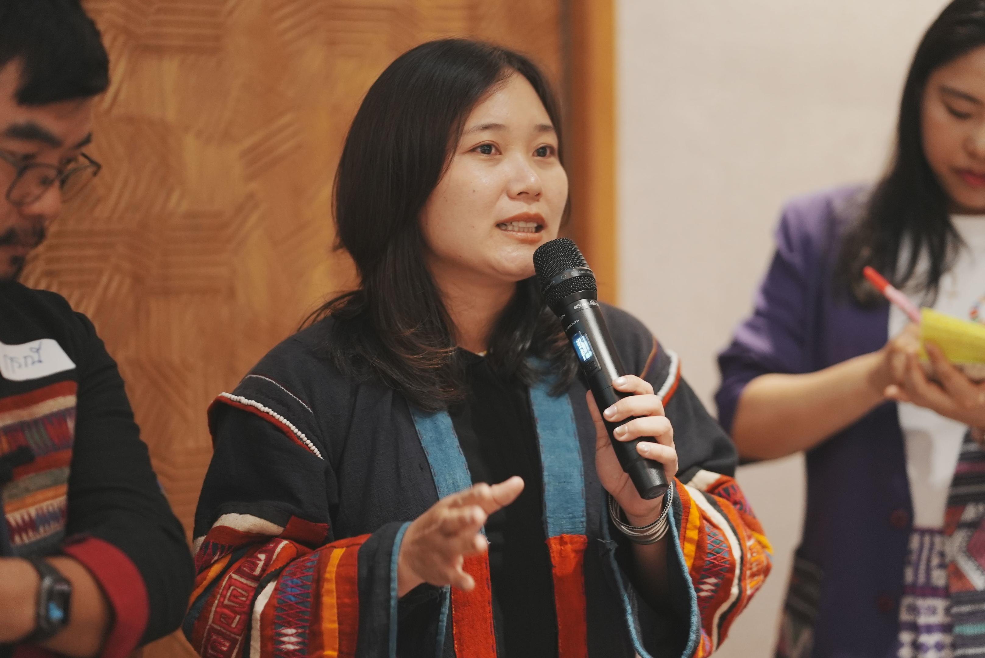 Woman in traditional clothing speaks into a microphone while others listen.