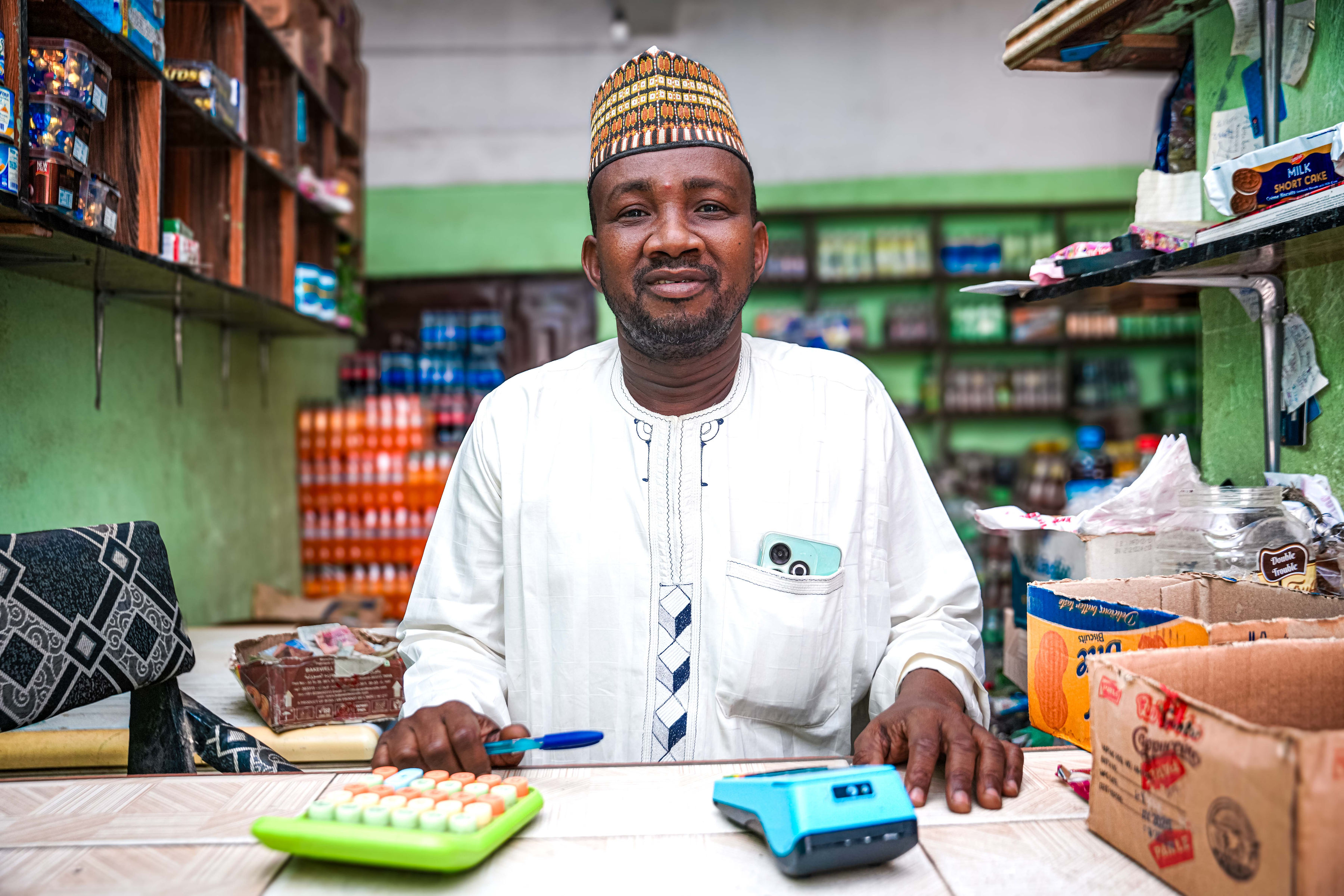 A smiling man in traditional attire sits behind a shop counter with items and shelves in the background.
