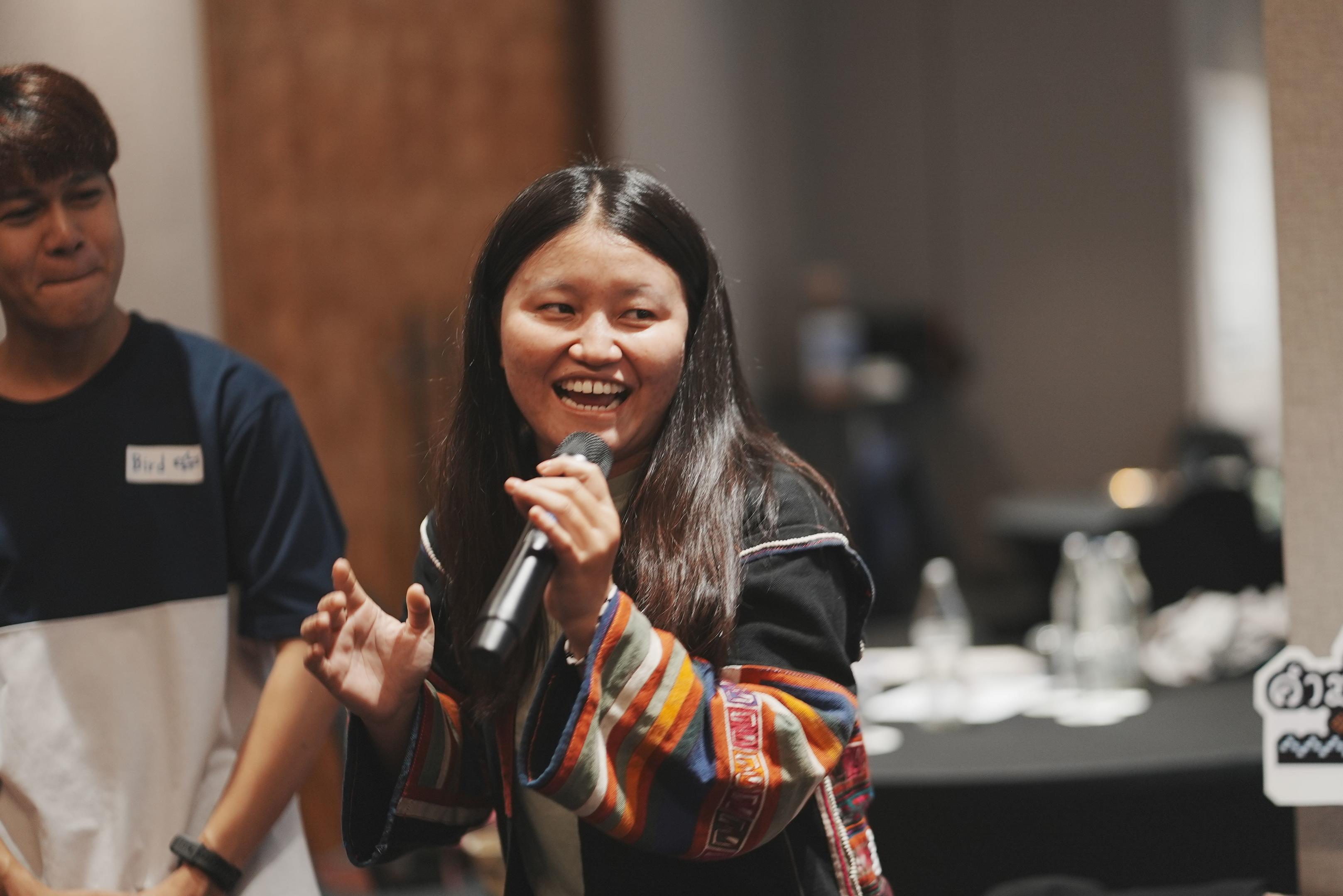 Woman smiling and singing into a microphone, with a man smiling in the background.