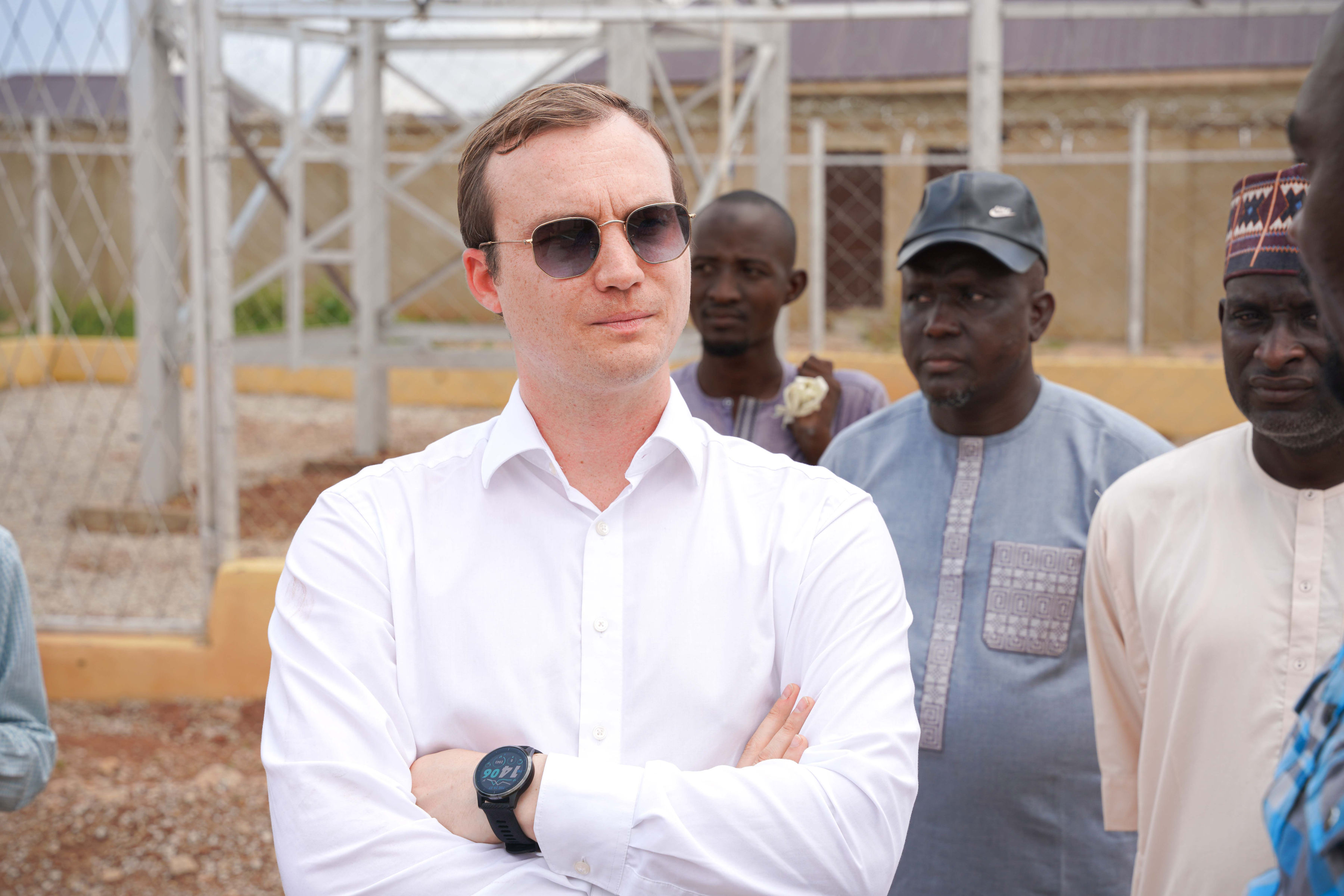 A man in sunglasses stands with crossed arms, surrounded by a group of men in a rural setting.