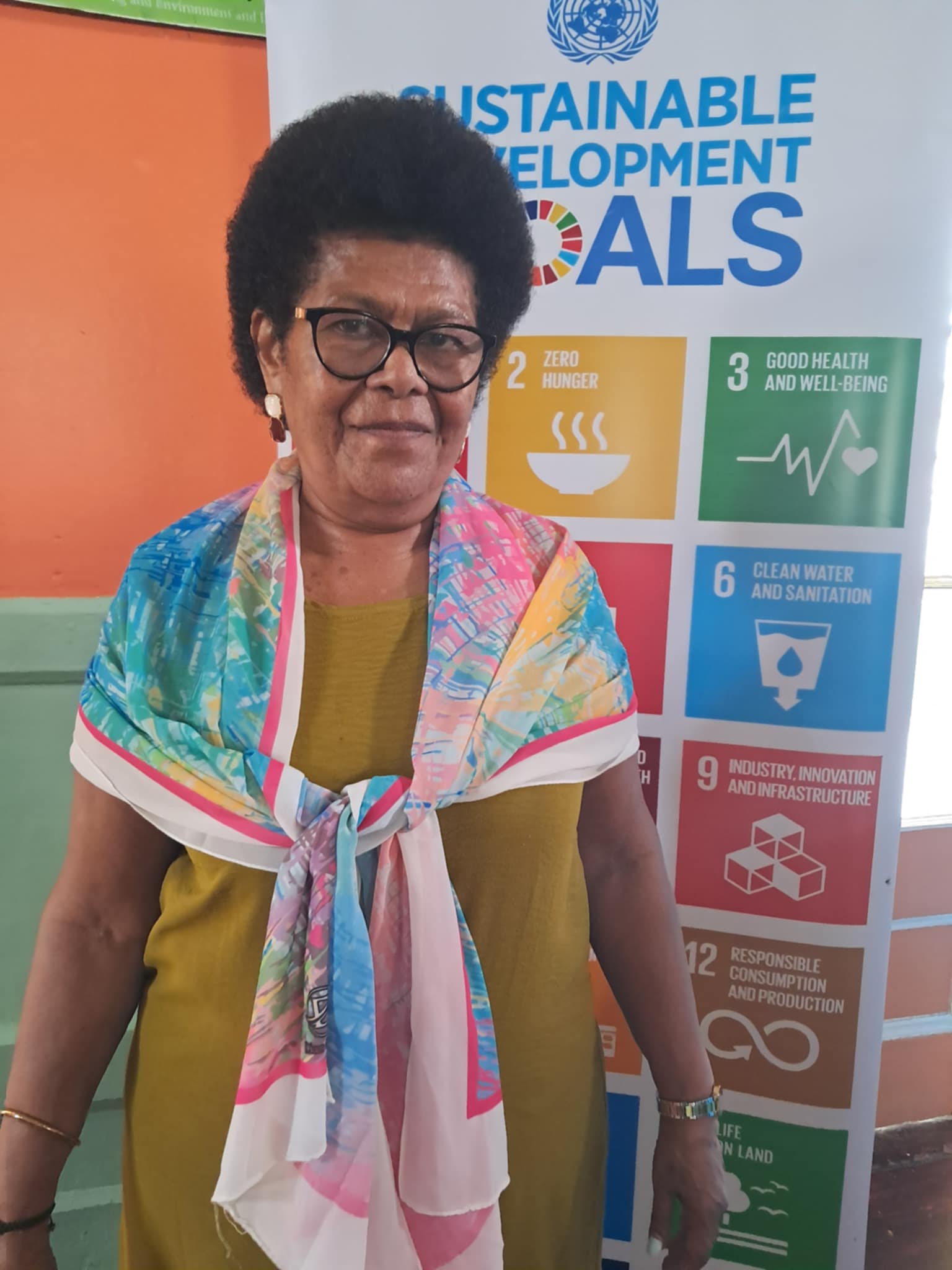Woman with glasses wearing a colorful scarf, standing in front of a Sustainable Development Goals backdrop.