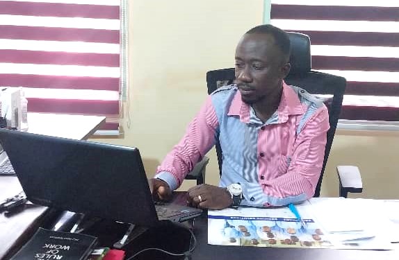 A man working at a desk with a laptop, wearing a pink and blue shirt, focused on his task.