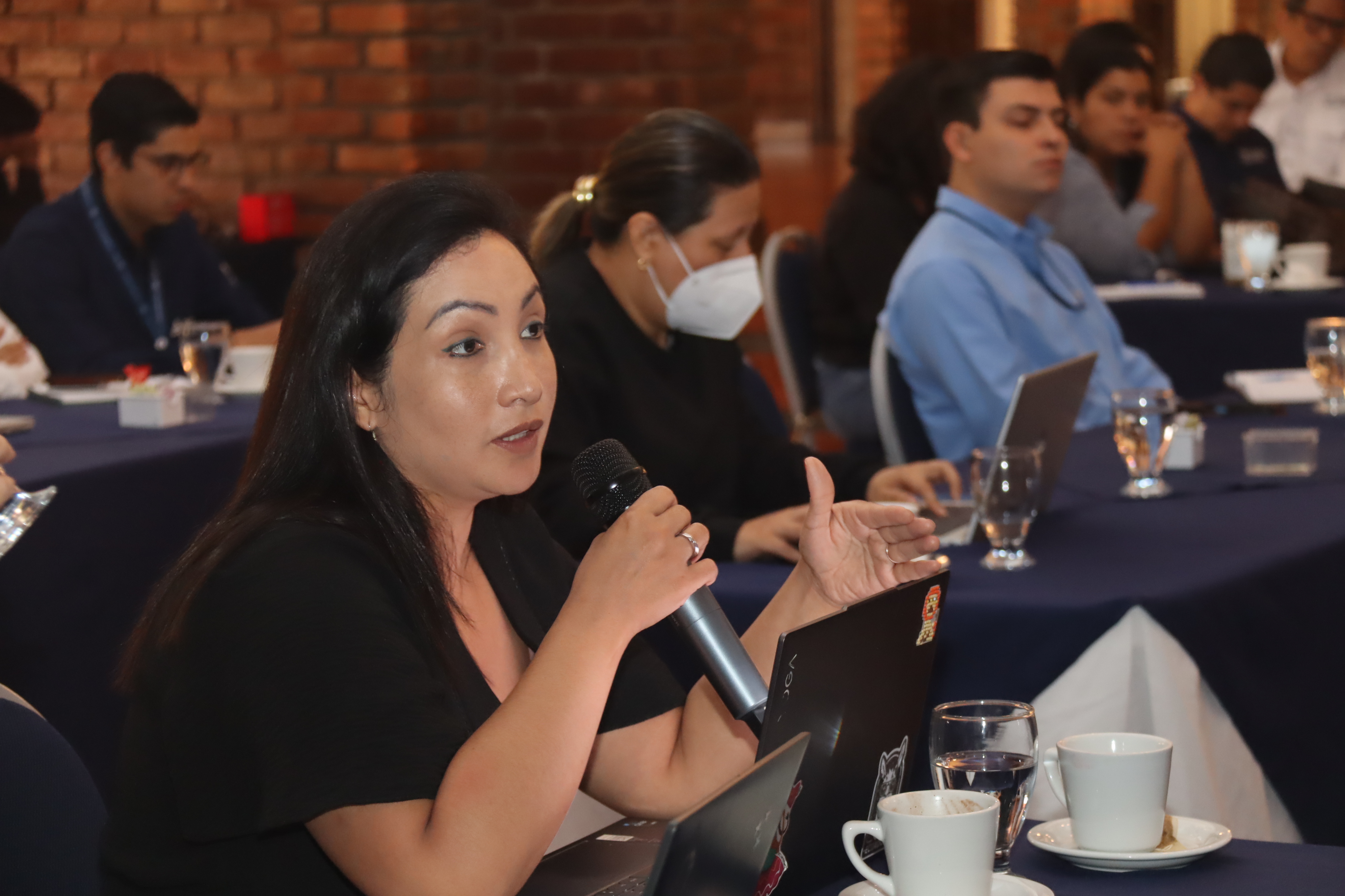 A woman speaks into a microphone during a conference, with attendees engaged in the background.