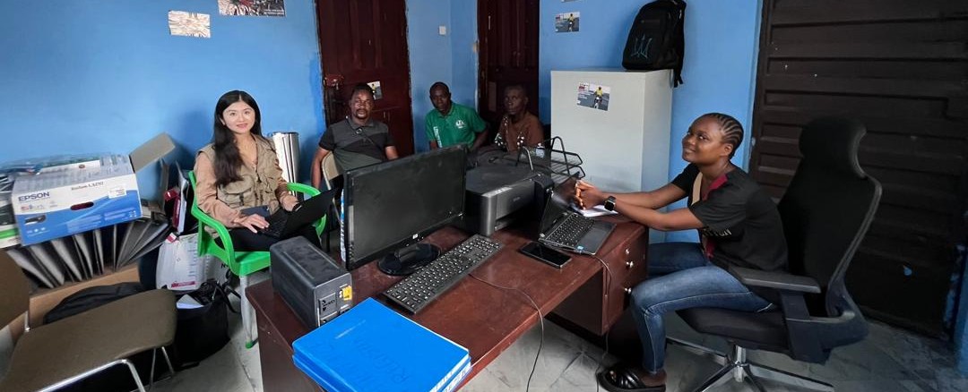 A diverse group of five people sits in a casual workspace with computers and office supplies.
