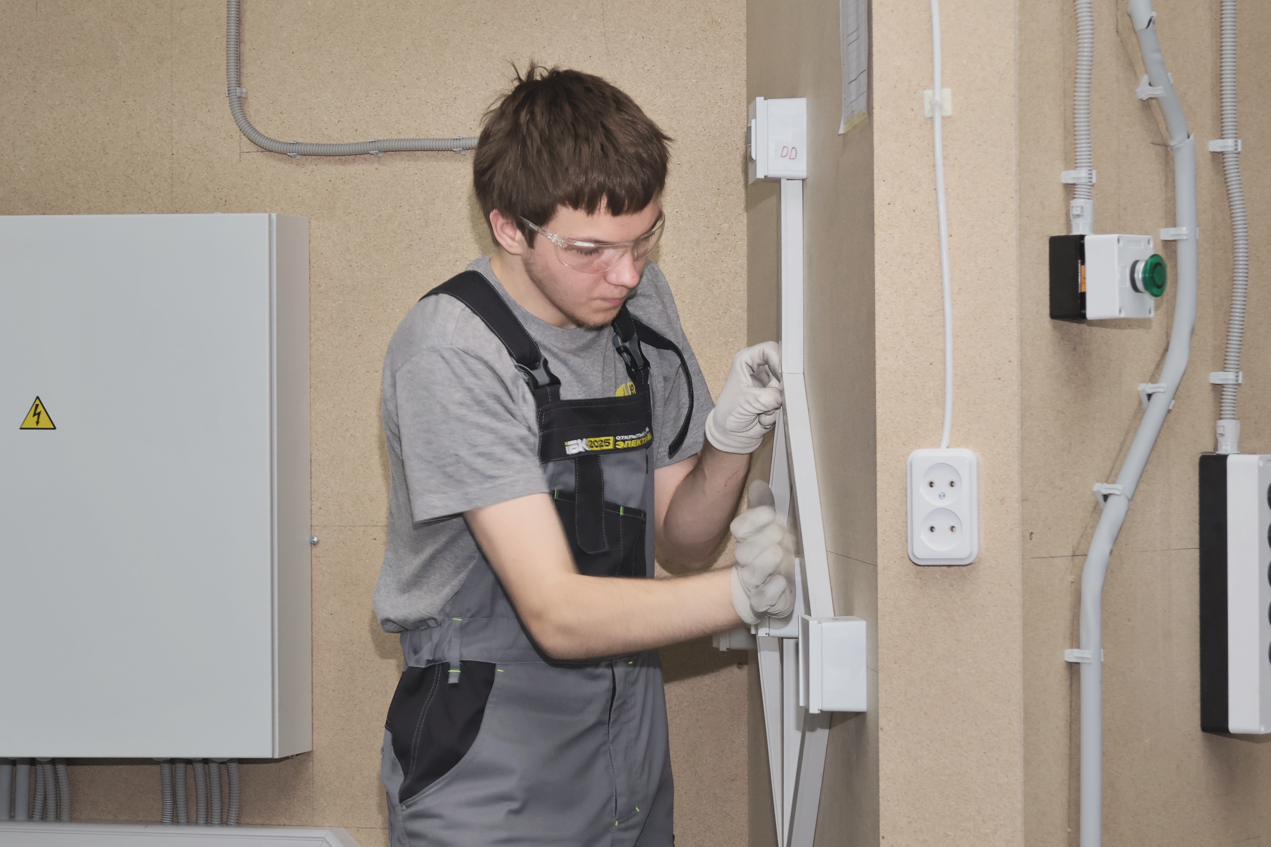 A young technician in grey overalls installs electrical wiring on a wall.