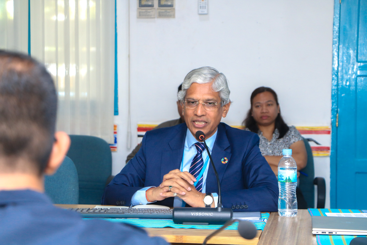 A man in a suit speaks at a conference table, while a woman sits in the background.