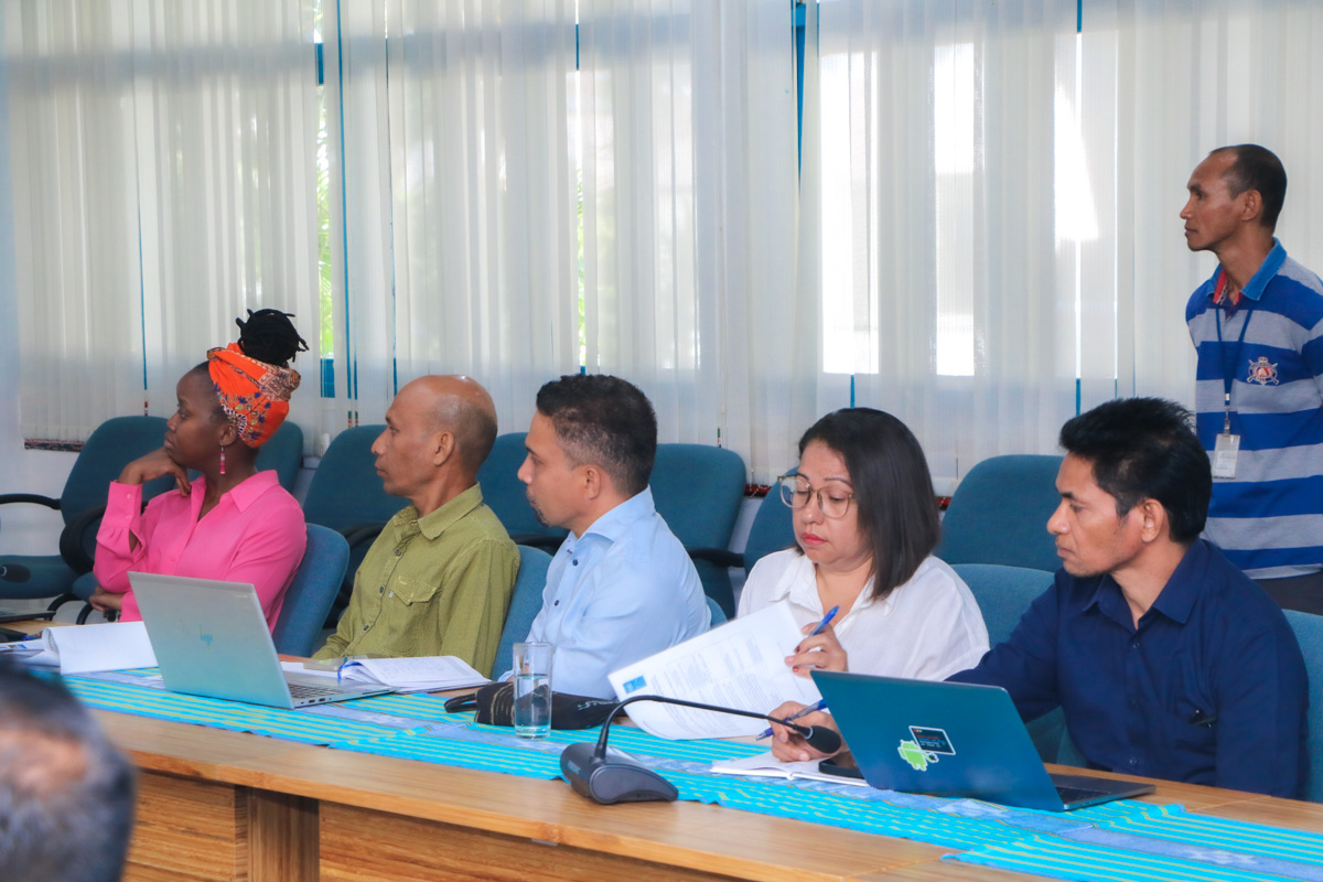 A group of five people seated around a table, discussing with laptops in a bright room.