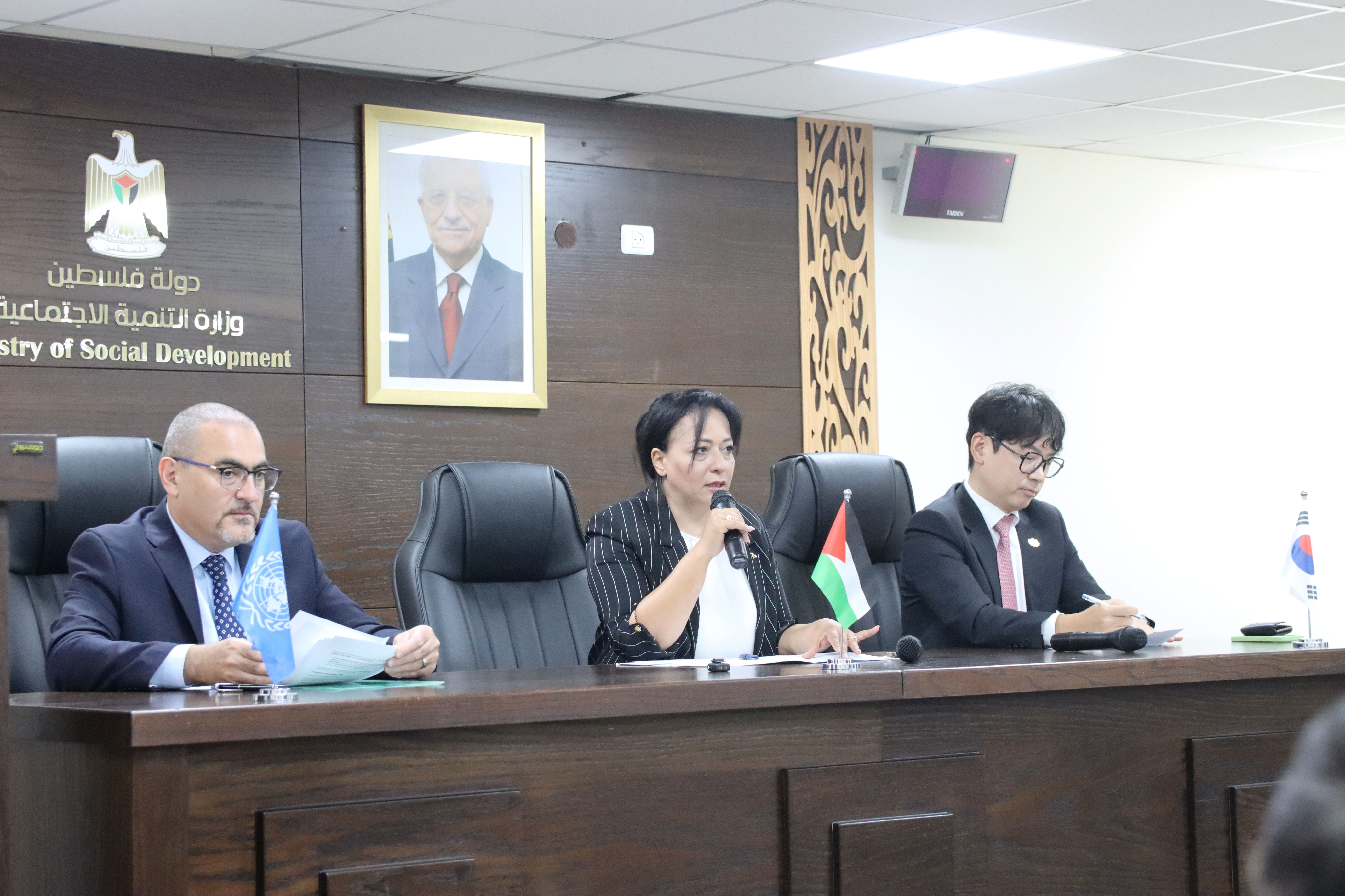Three officials seated at a table, one speaking, with flags and a portrait in the background.