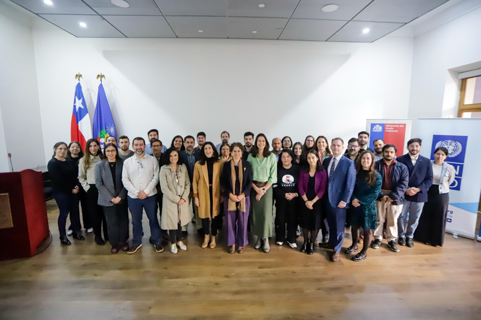 Group photo of diverse individuals standing together in a well-lit room with flags in the background.