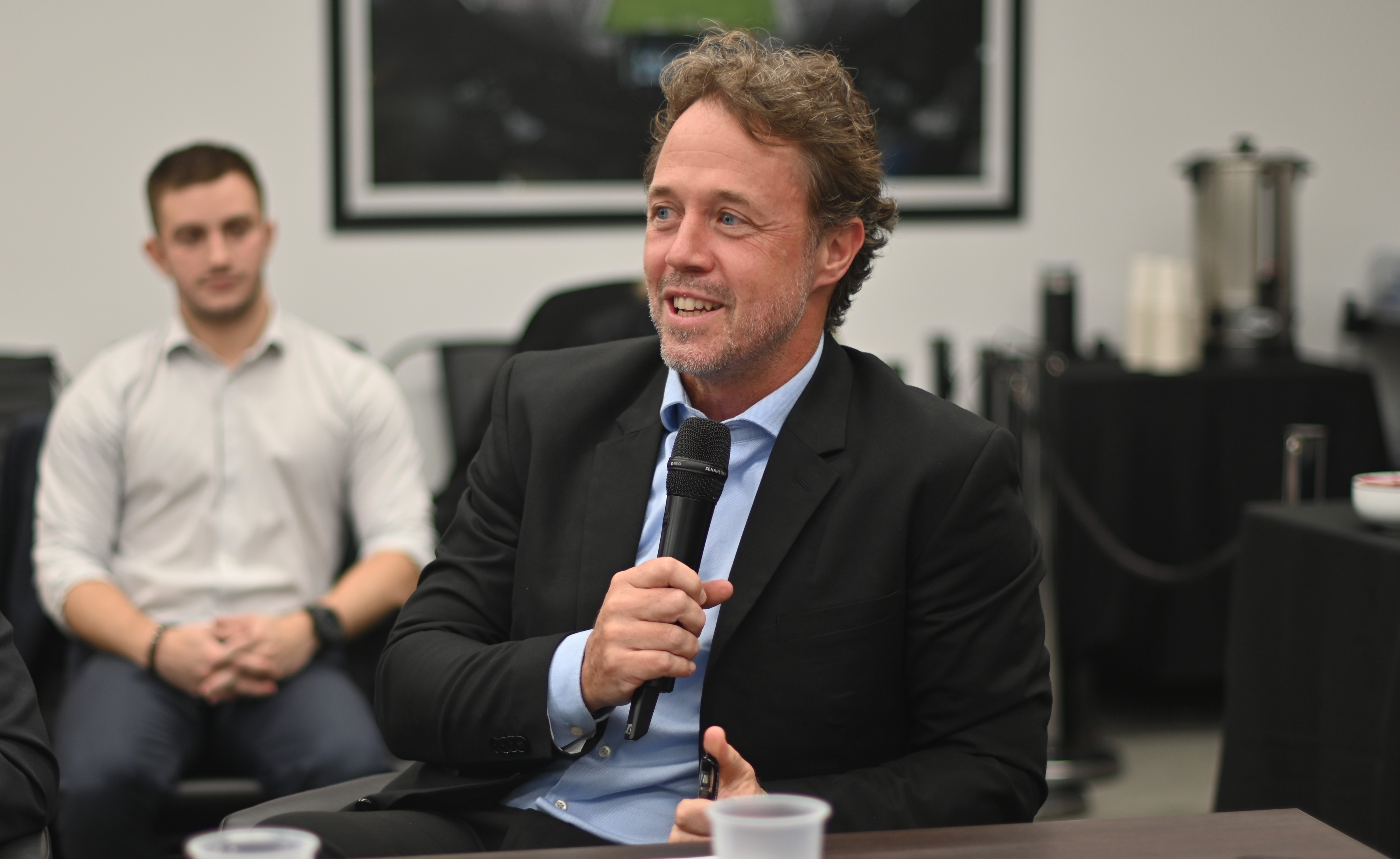 Un hombre con un traje negro habla con un micrófono mientras otro hombre escucha en el fondo.