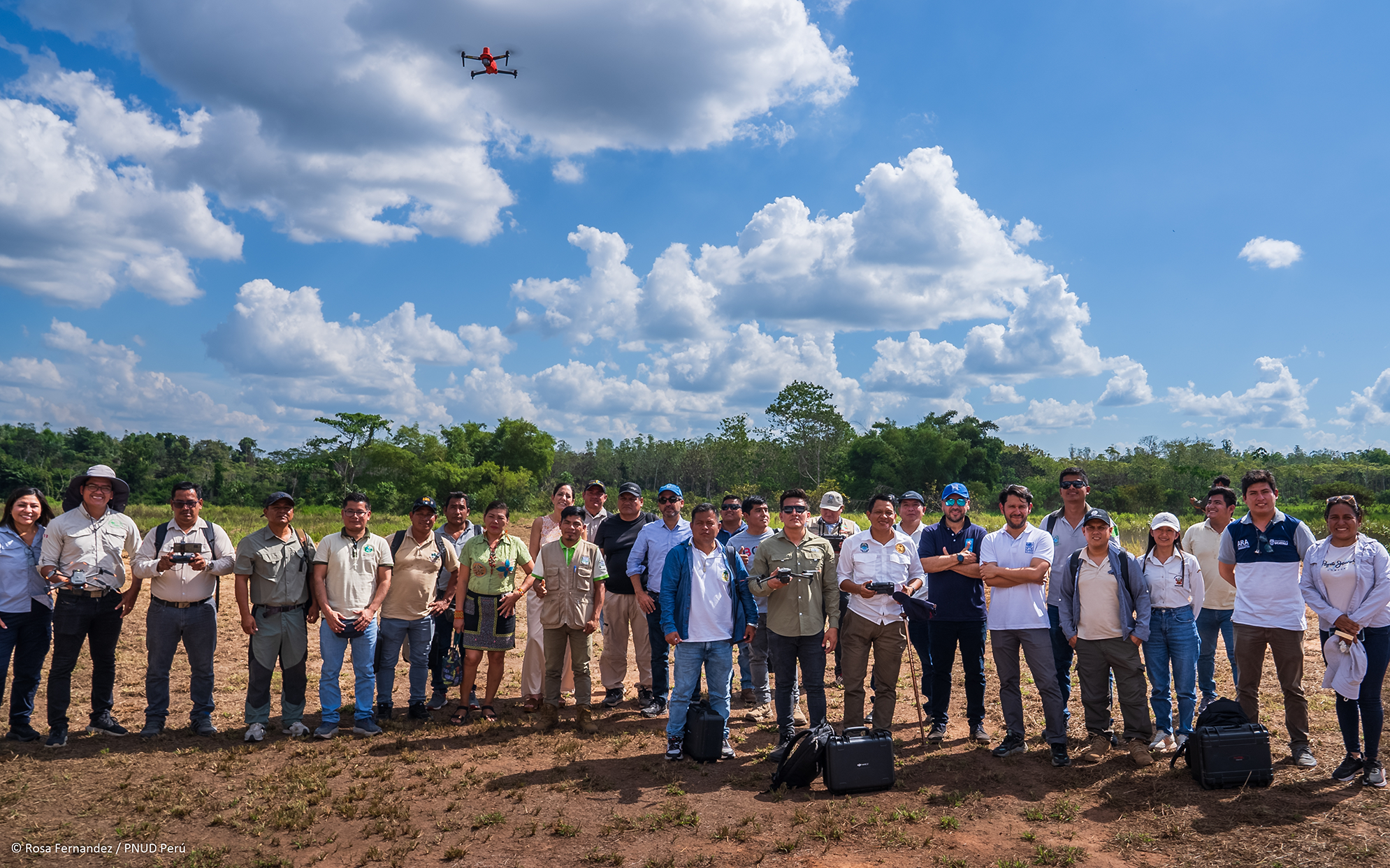 A group of diverse individuals stands in an open field under a blue sky with clouds, a drone flying above.