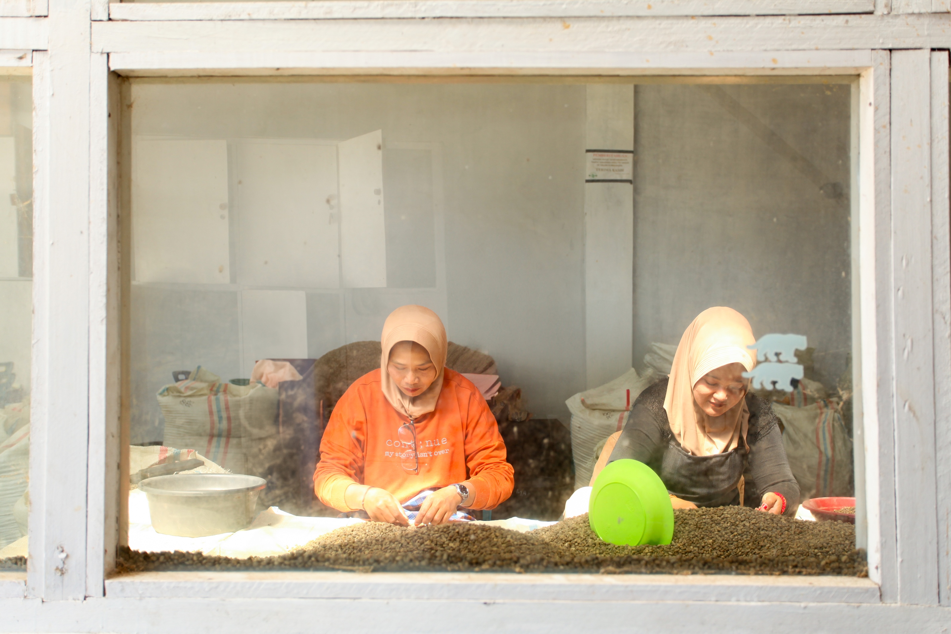Two women sorting tea leaves behind a glass window, one in orange and one in black.