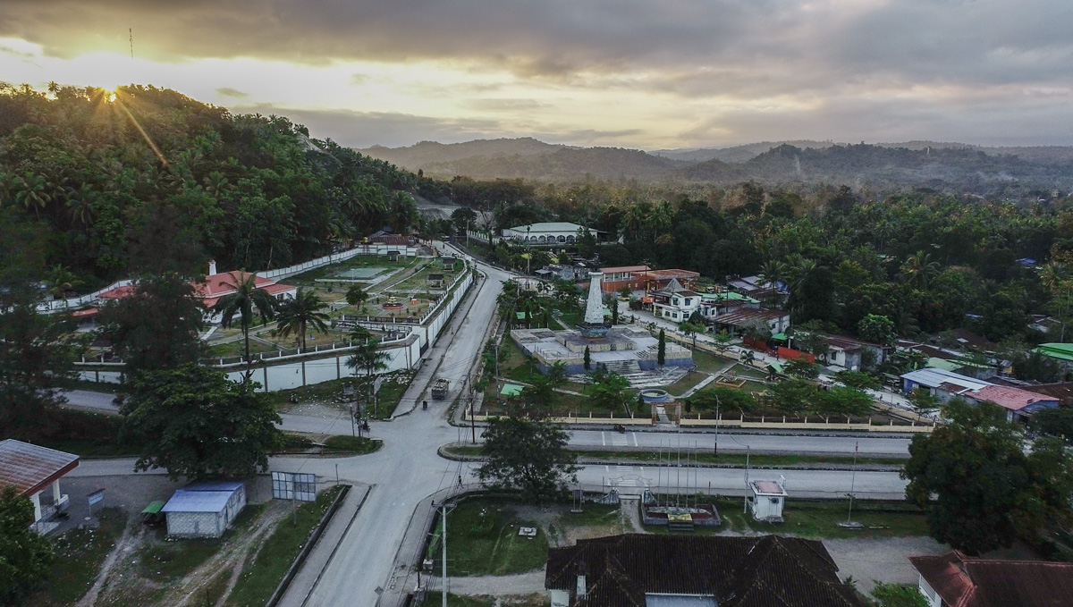 Aerial view of a serene landscape with roads, greenery, and a distant sunset over hills.