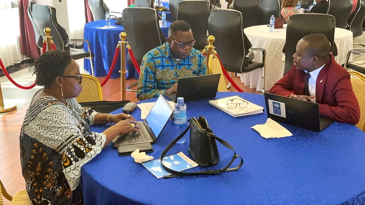 Three people seated at a blue table, working on laptops in a conference setting.