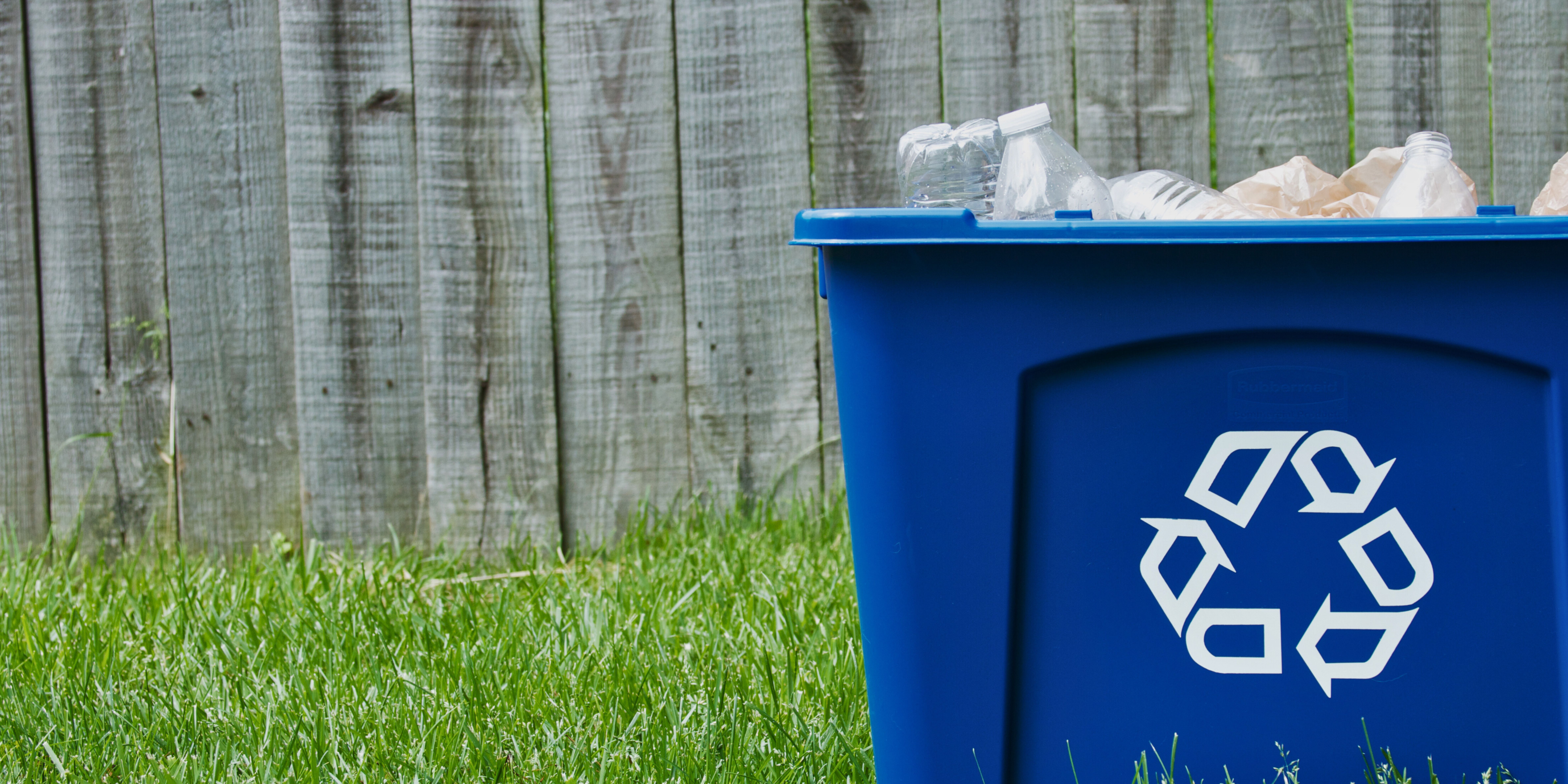 A blue recycling bin filled with recyclables, set against a wooden fence and grass.