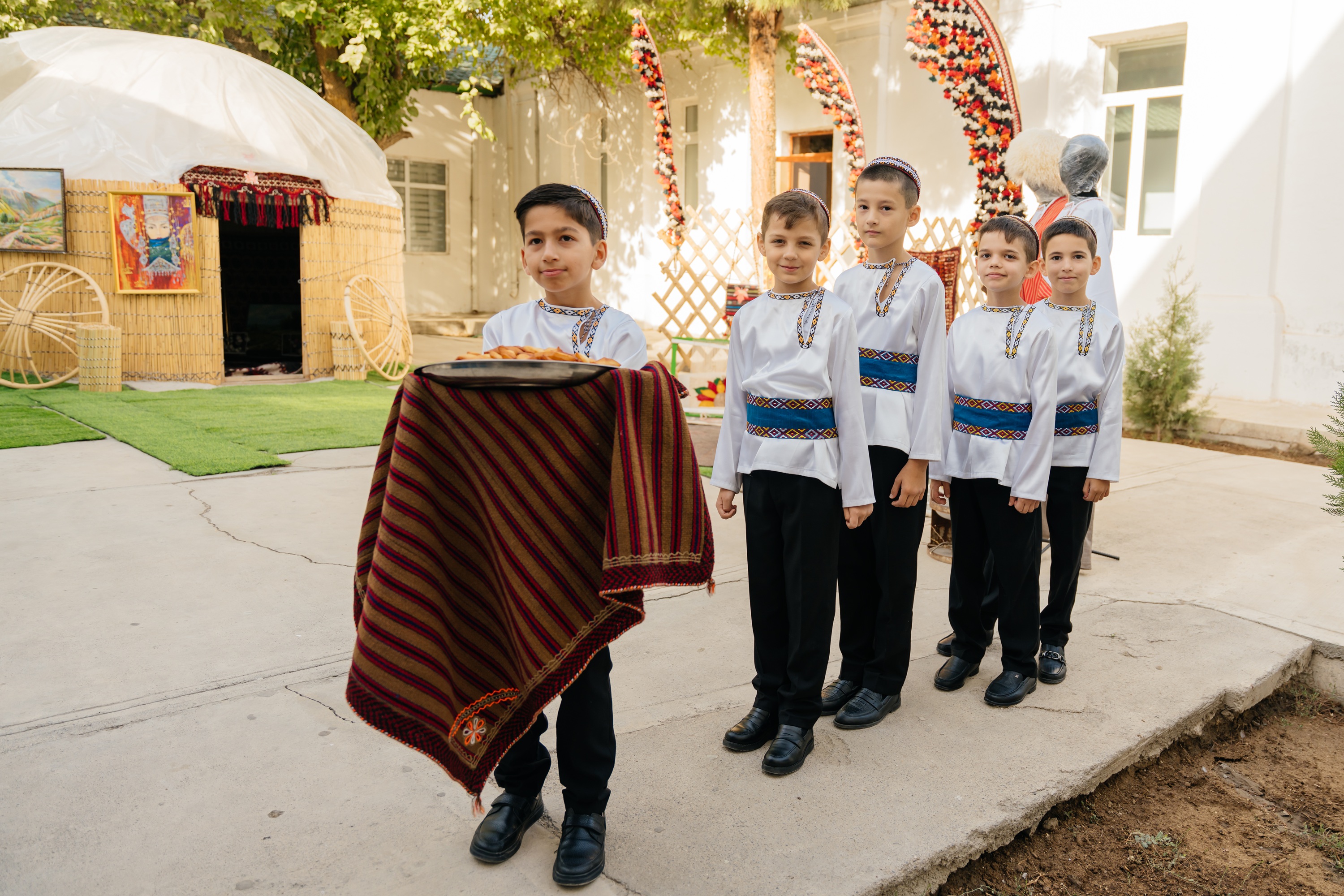 Boy in striped poncho leads a line of children in white shirts in a sunny courtyard.