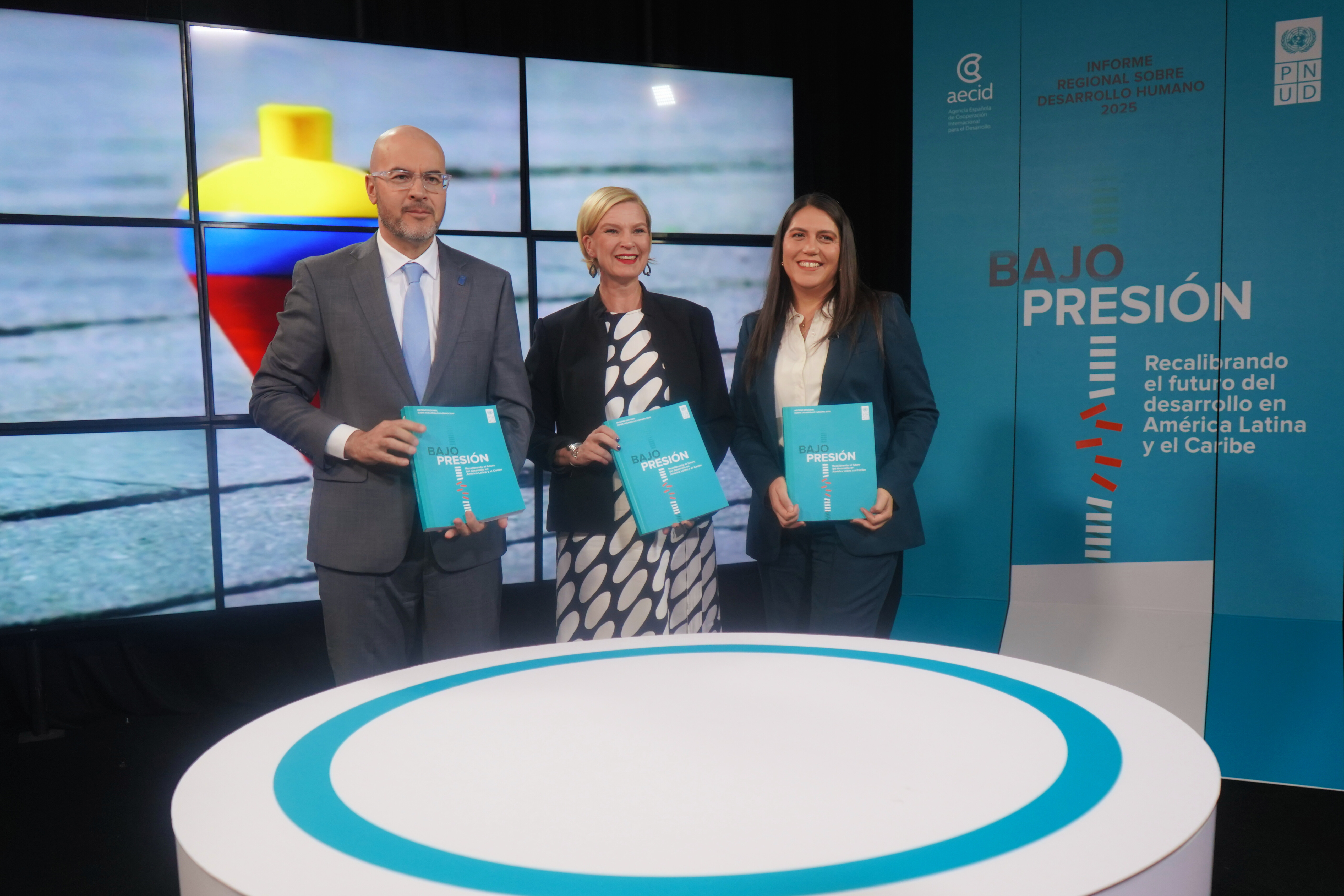 Three presenters in a blue studio hold teal booklets; circular stage and a screen displaying a flag.