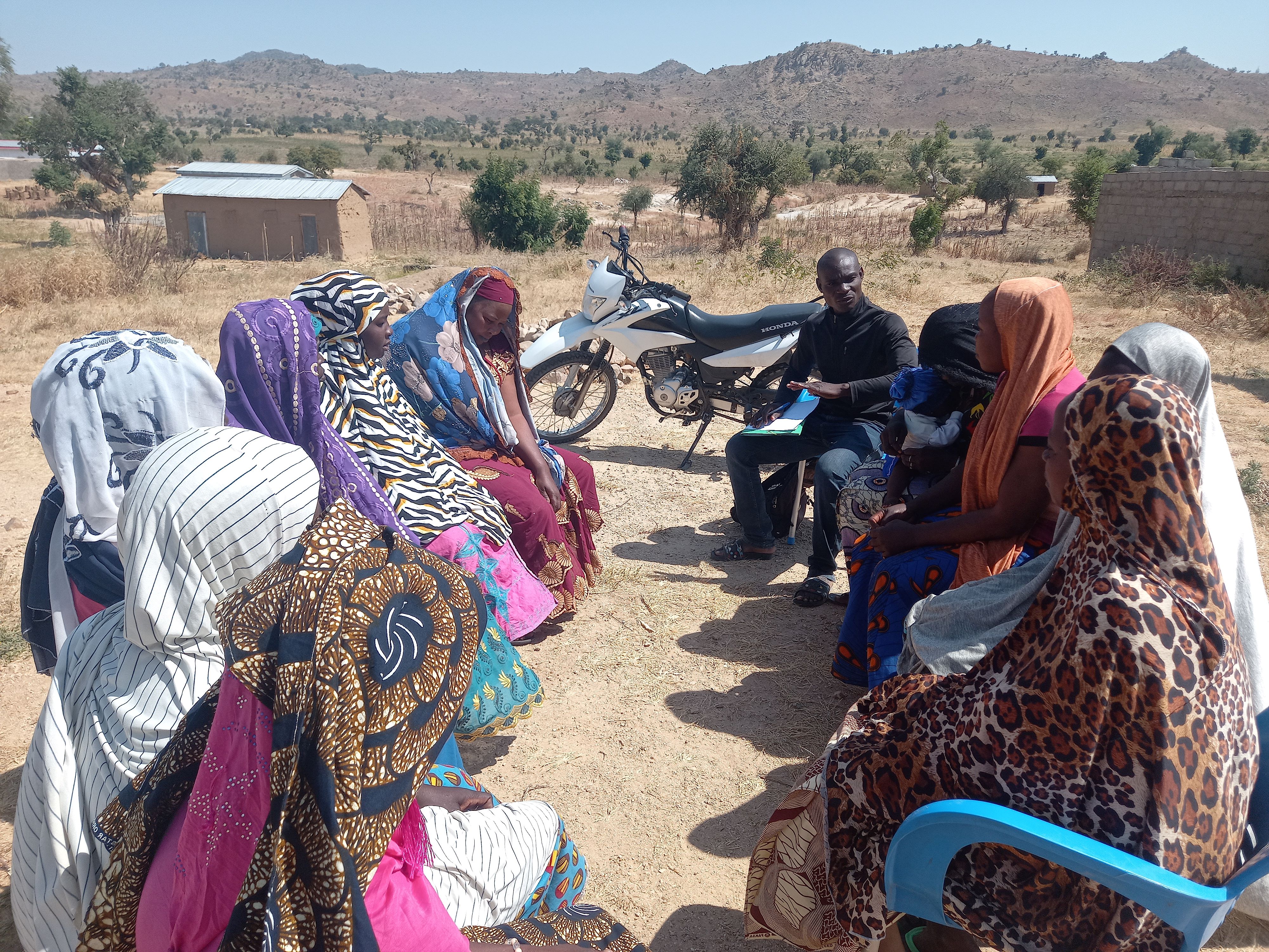 Group of women in colorful hijabs sit in a circle outdoors, mountains in the background.