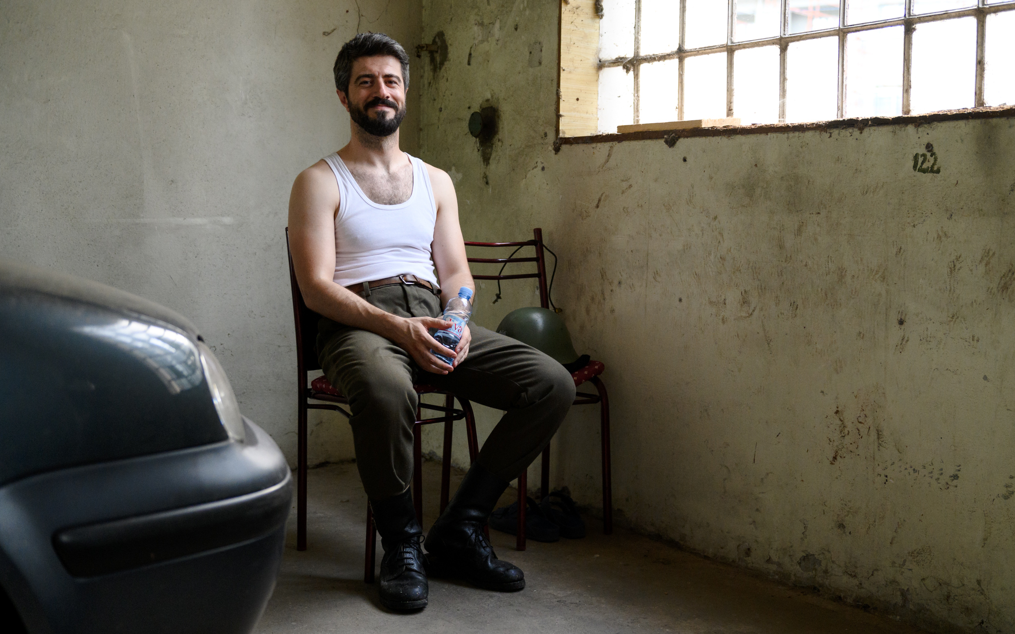Bearded man in a white tank top sits on a chair beside a car in a dim, worn room with a window.