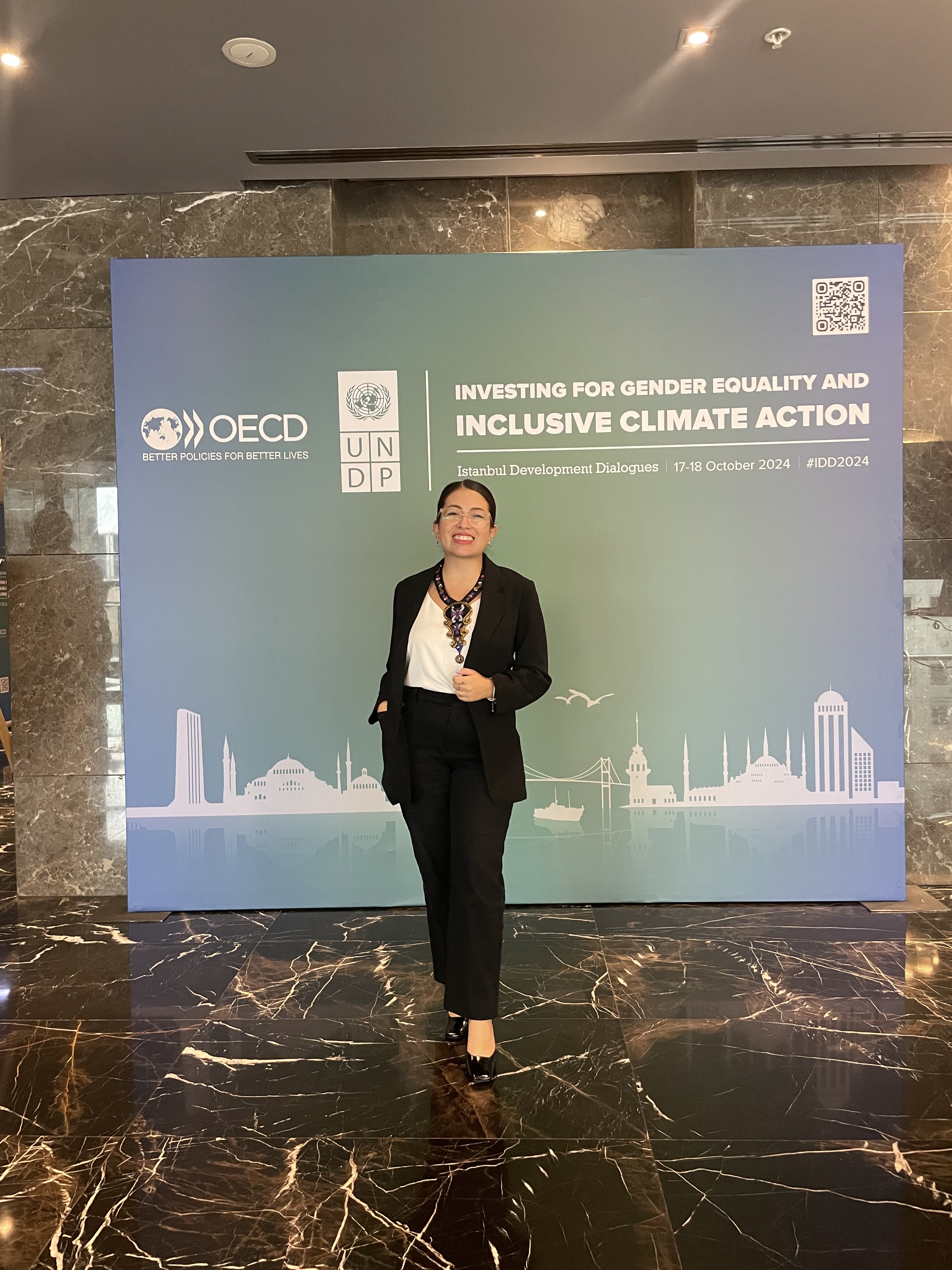 Photograph of a woman in a black suit standing in front of an OECD banner with a city skyline.