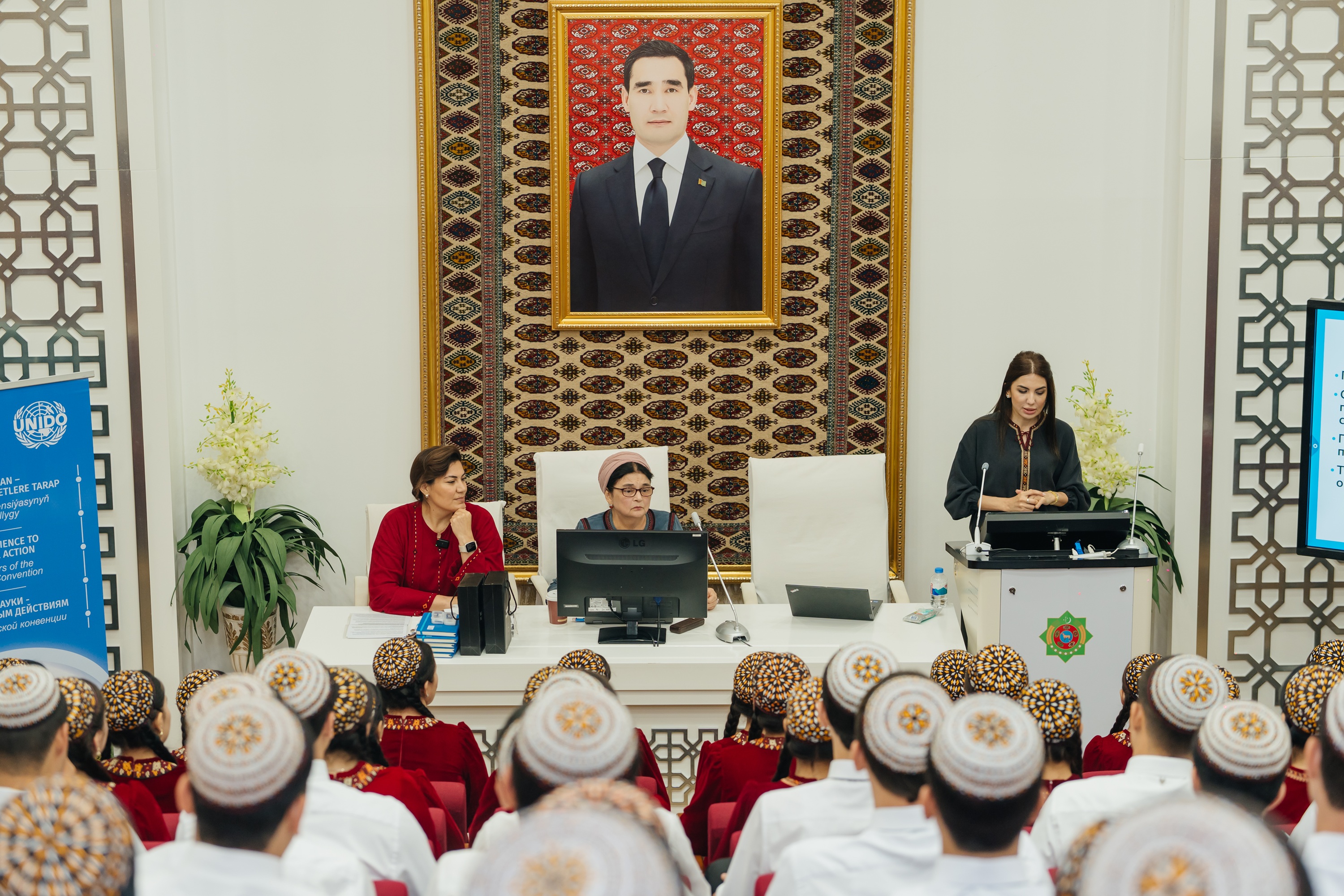 Audience in a ceremonial hall wearing white caps, with speakers on stage and a framed portrait of a man on the wall.