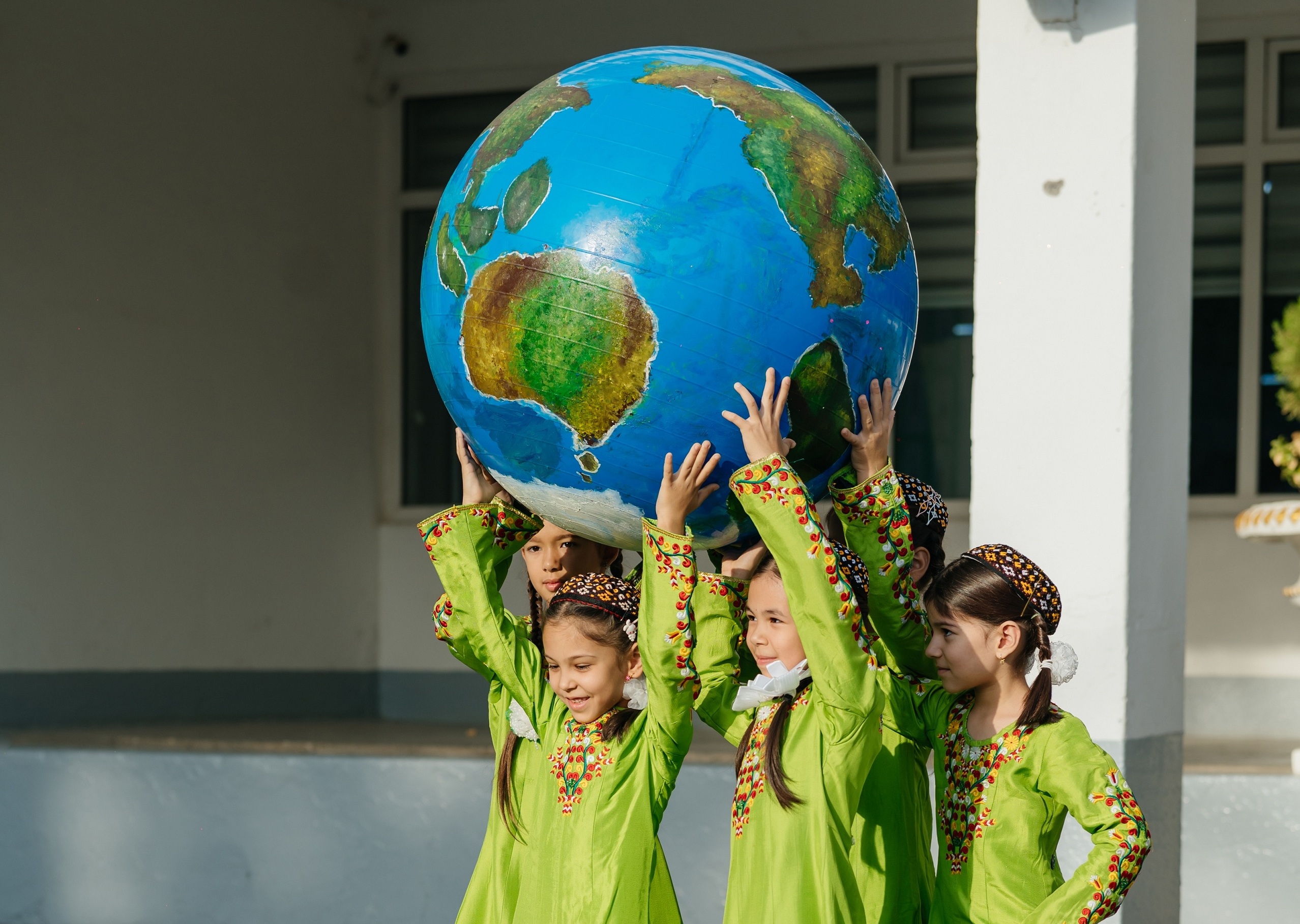 Three smiling children in lime shirts lift a large globe outdoors in a schoolyard.