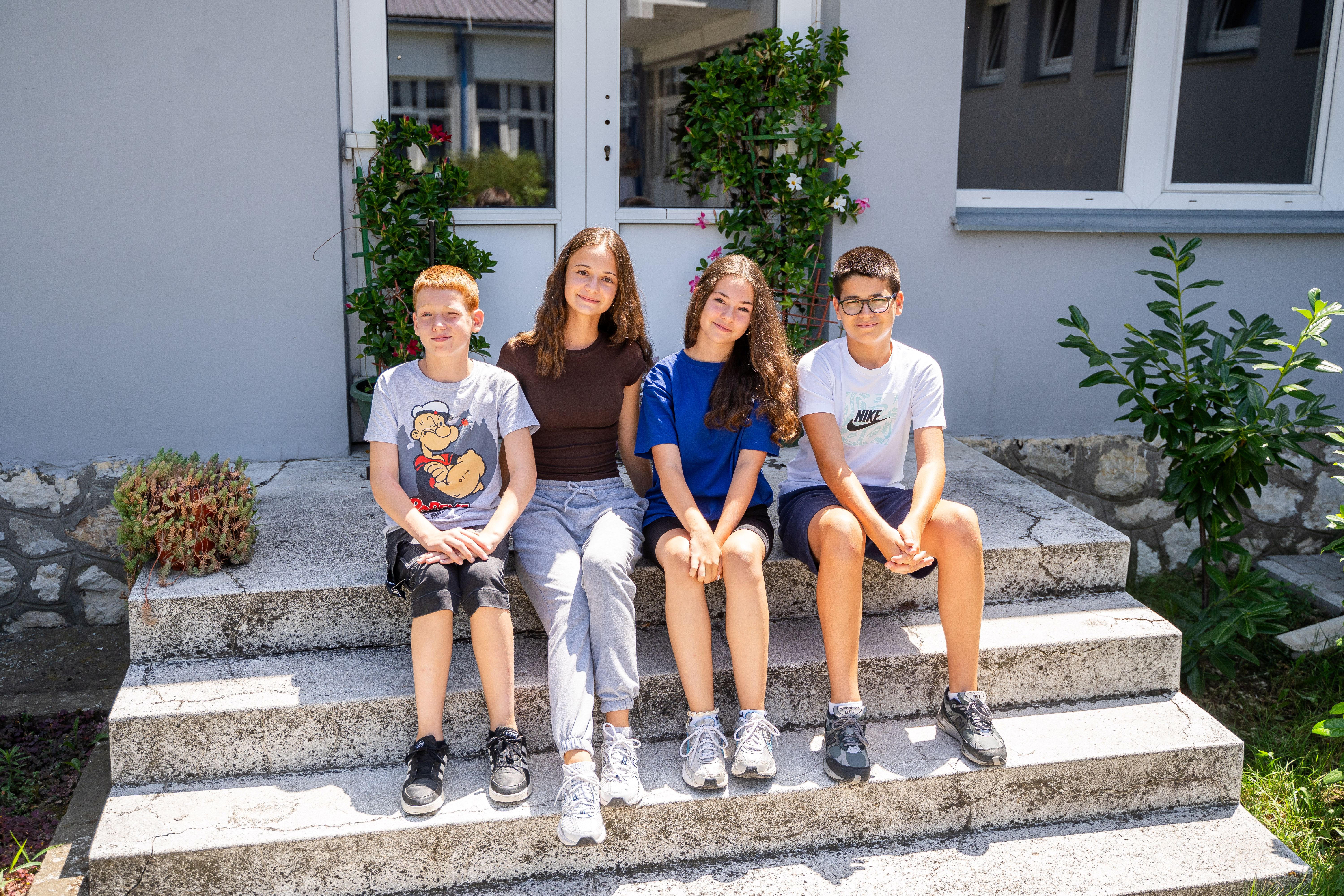 Five teens sitting on concrete steps outside a building, smiling.