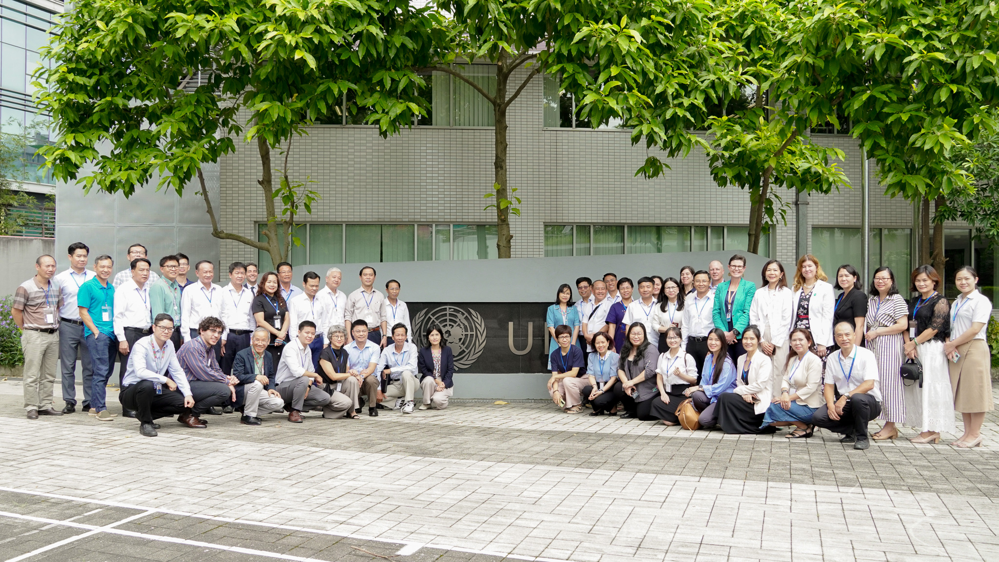 Large group of people posing for a photo outdoors in front of a light building with trees.