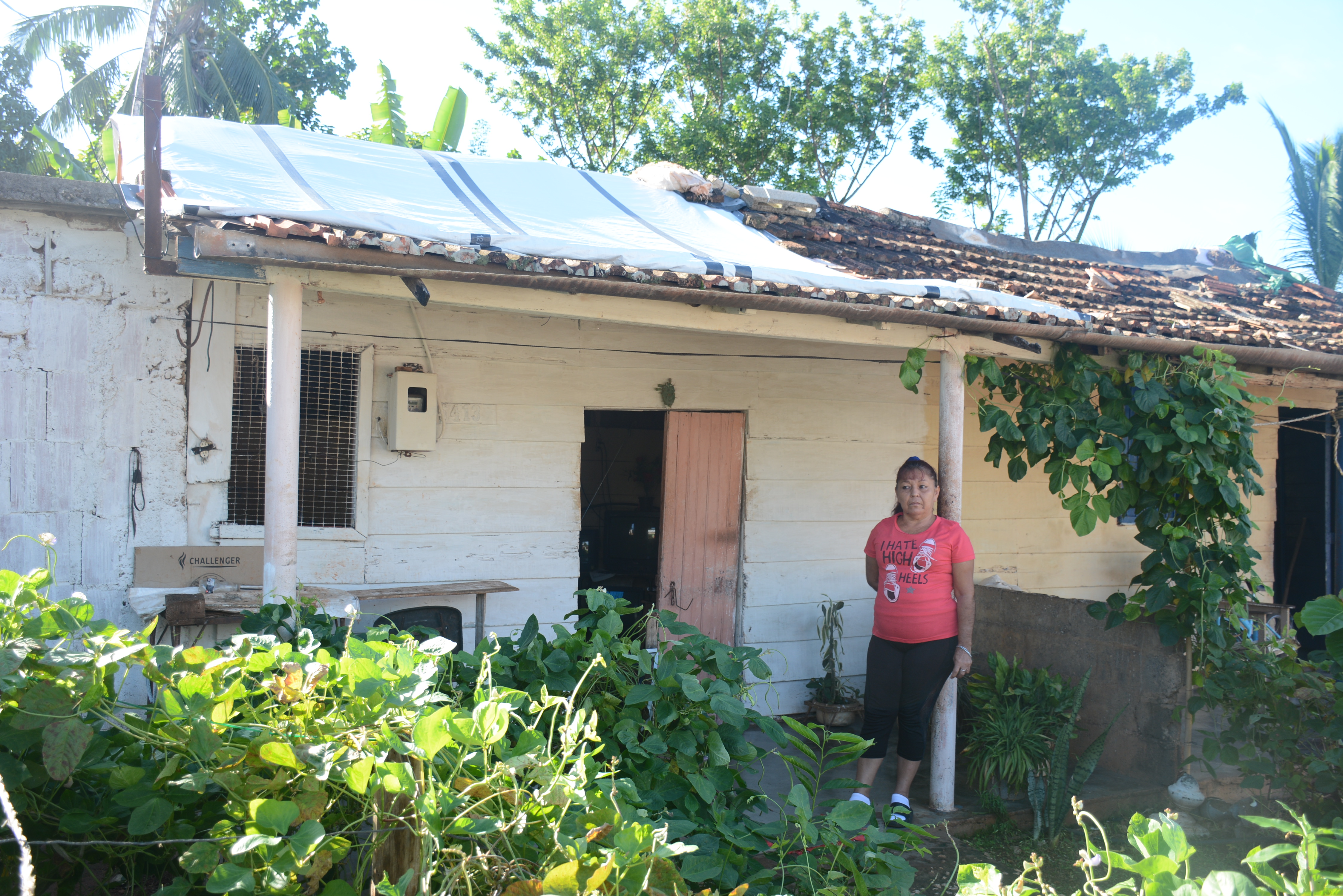 Photograph of a woman in a pink top standing outside a weathered cottage with a metal roof.