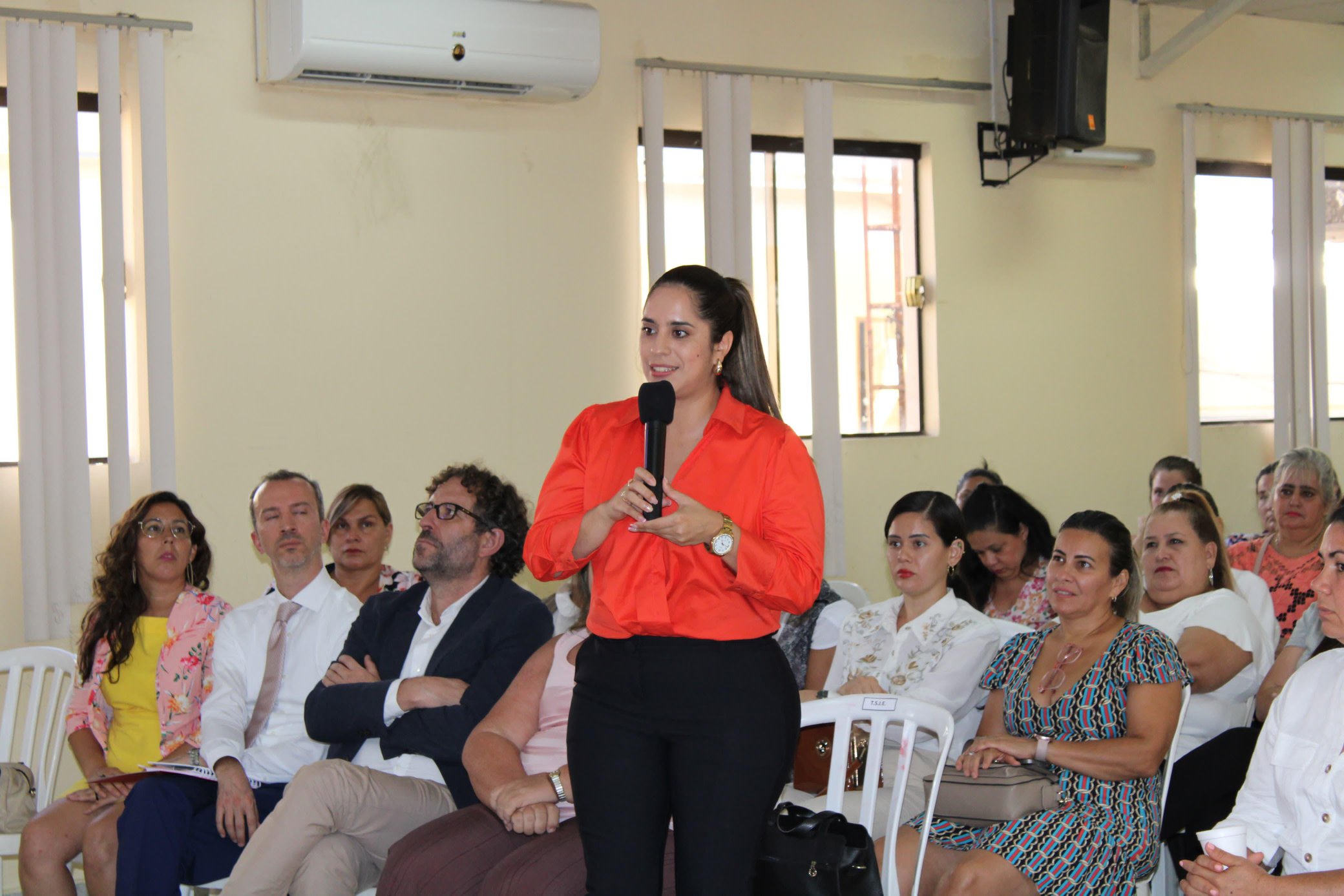 Woman in red blouse speaks into a microphone before an attentive audience in a conference room.