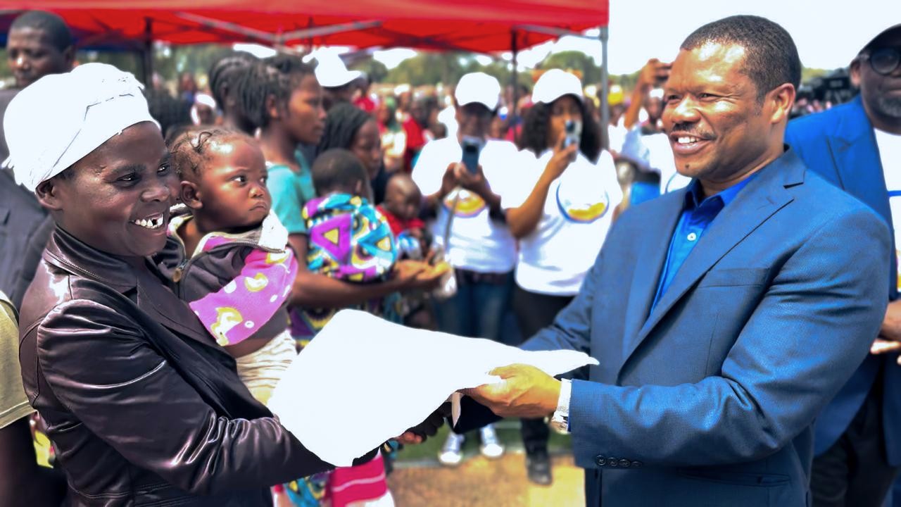 Man in blue suit hands a document to a woman under a red canopy at an outdoor ceremony.