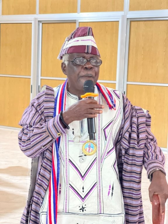 Person in colorful ceremonial regalia with purple detailing, holding a cup outdoors.