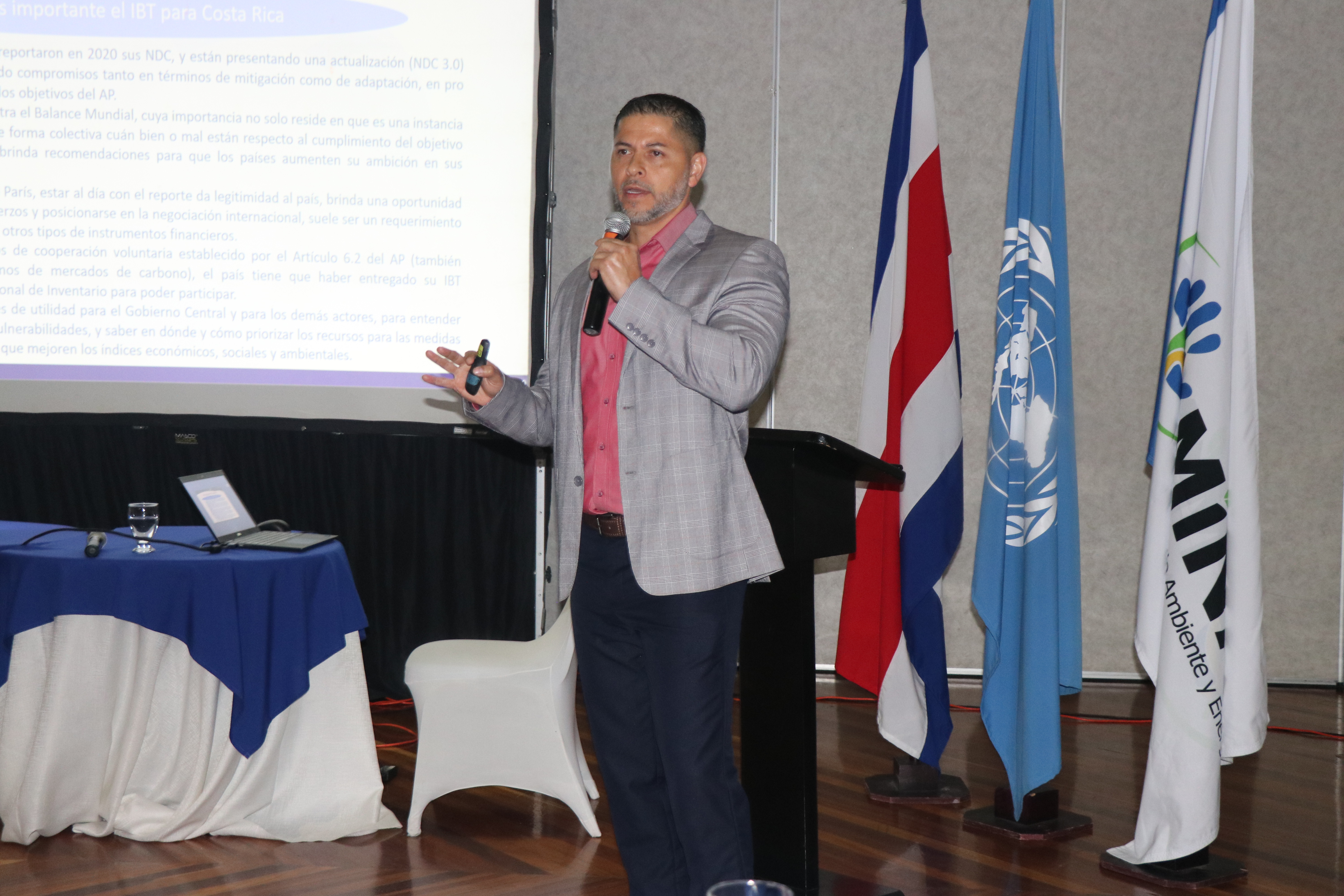 A speaker gestures while presenting at a conference, with flags in the background.