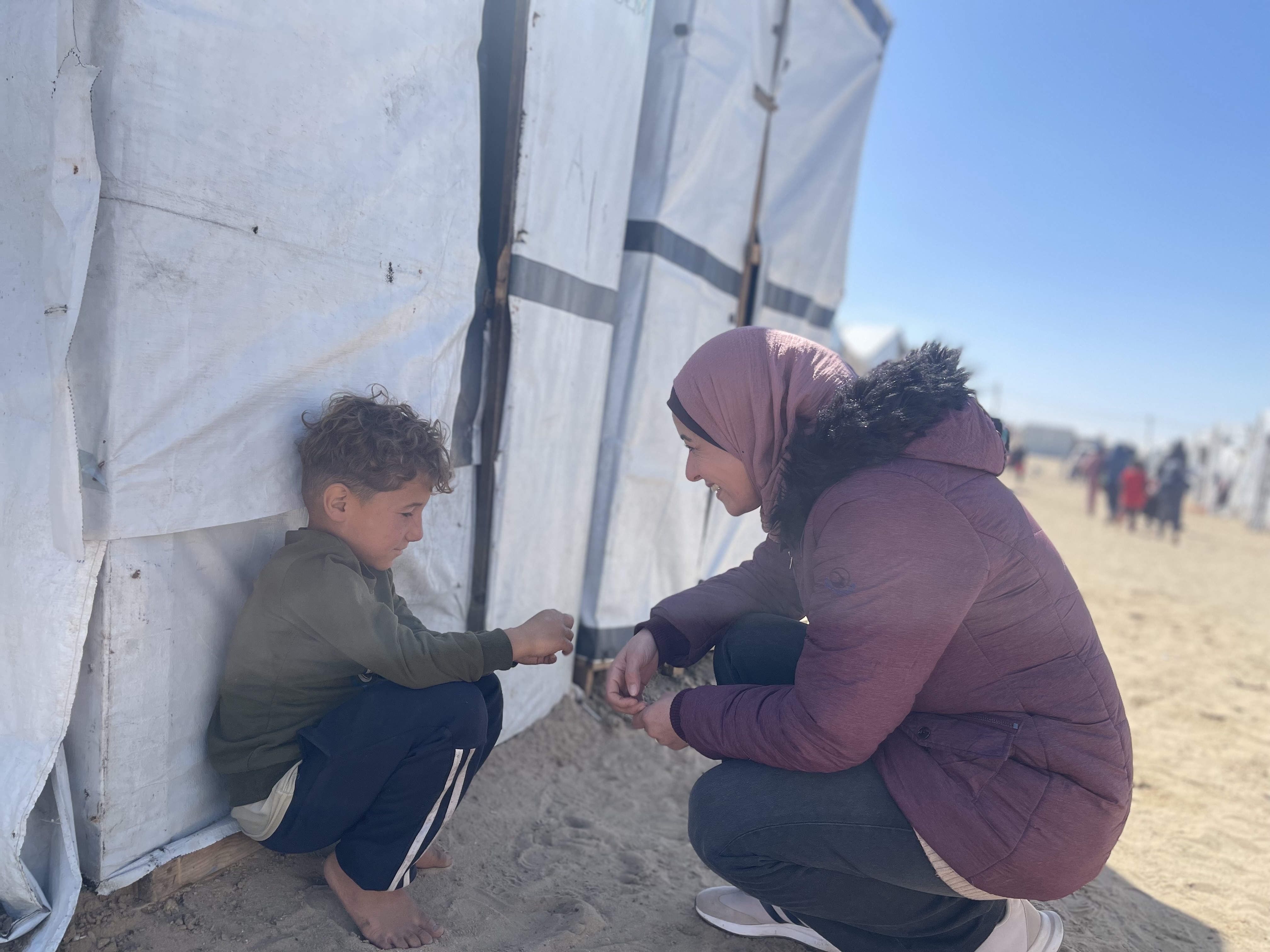 A woman in a purple jacket kneels beside a young boy near a tent in a refugee camp.