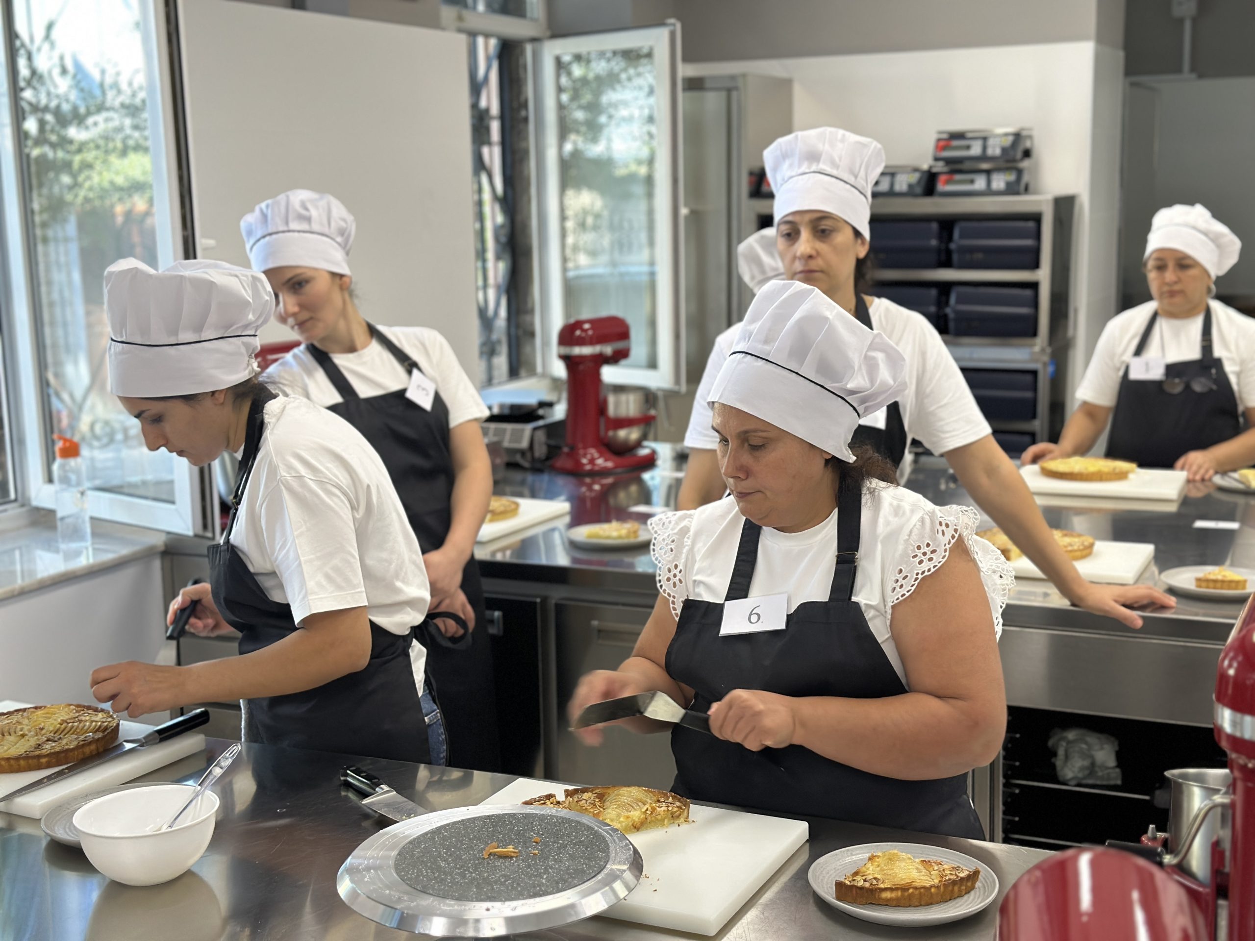Group of culinary students in white chef hats and aprons around a bright kitchen station.