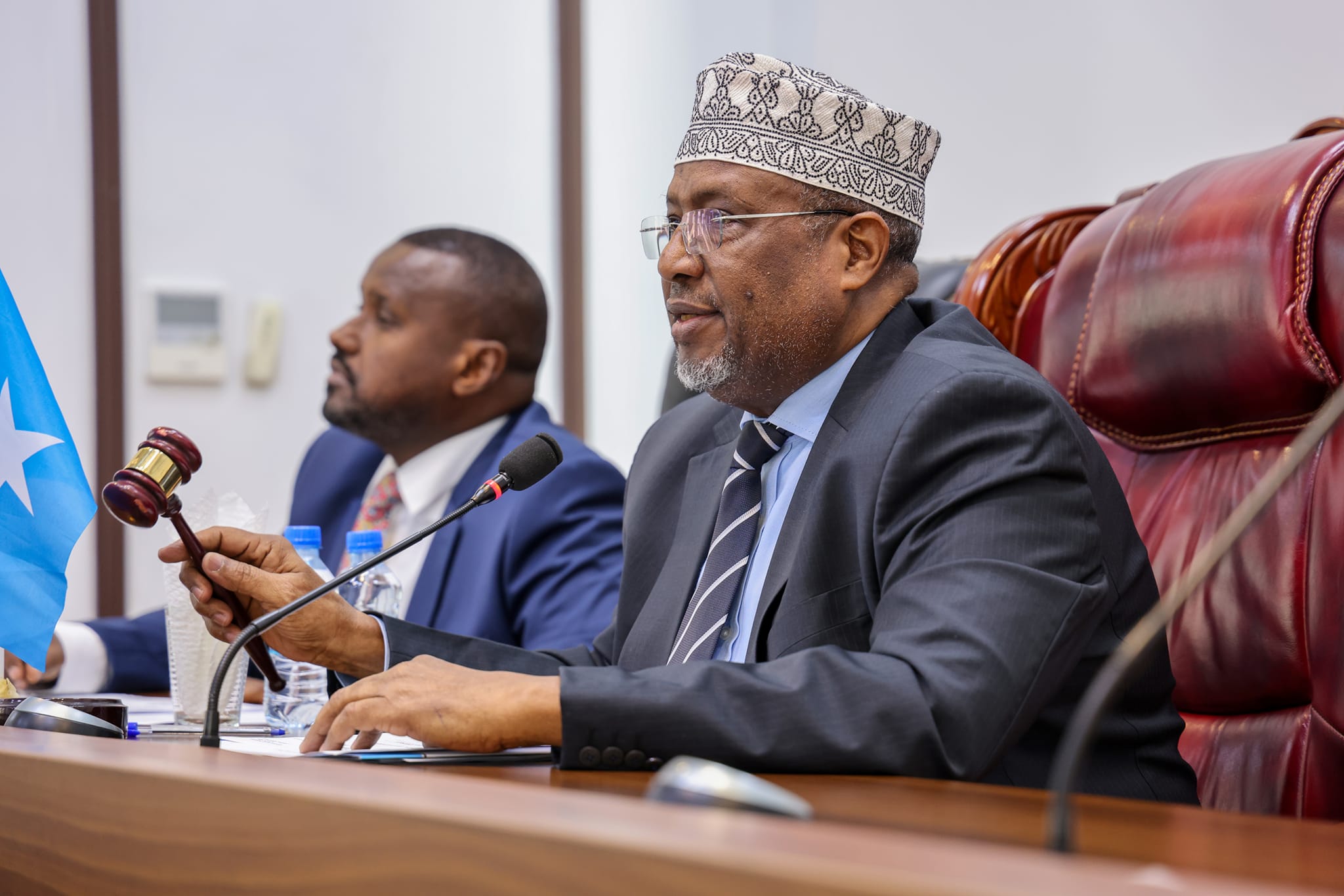 Crowned man in a suit at a council table with a microphone; blue flag with white star behind.
