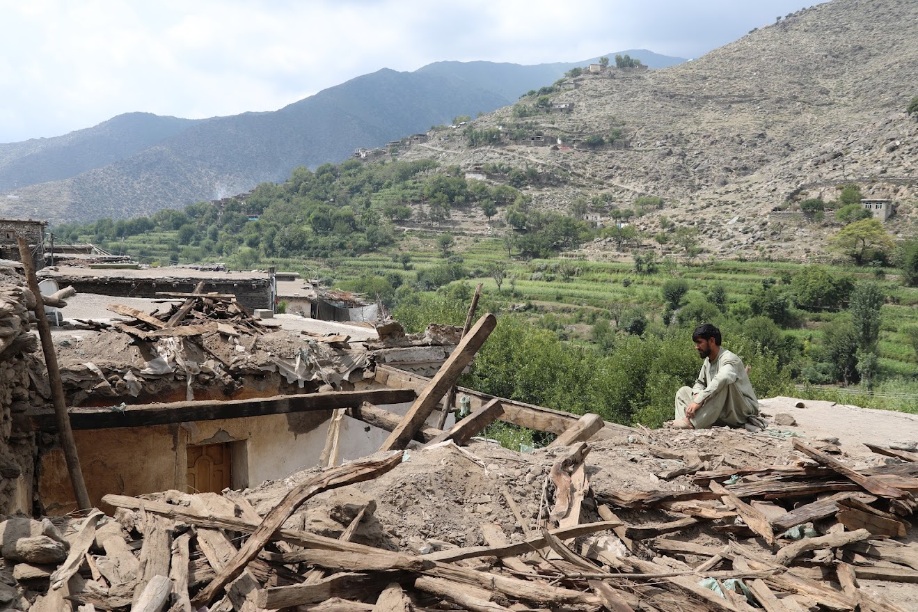Rubble and wooden debris in foreground; a person stands near a ruined structure with hills beyond.