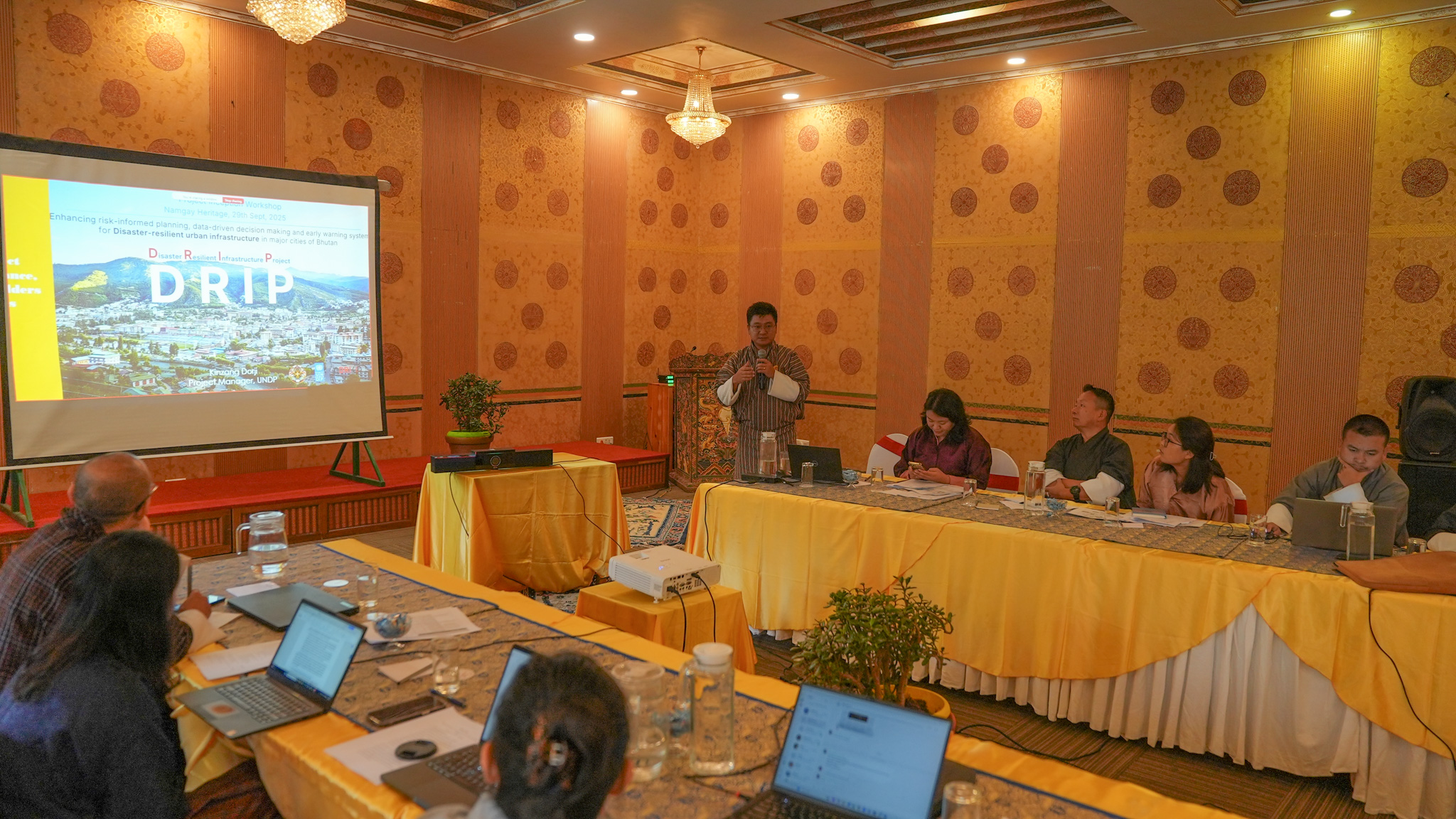 Photograph of panelists at a long table in a conference room, with a projector screen on the left.