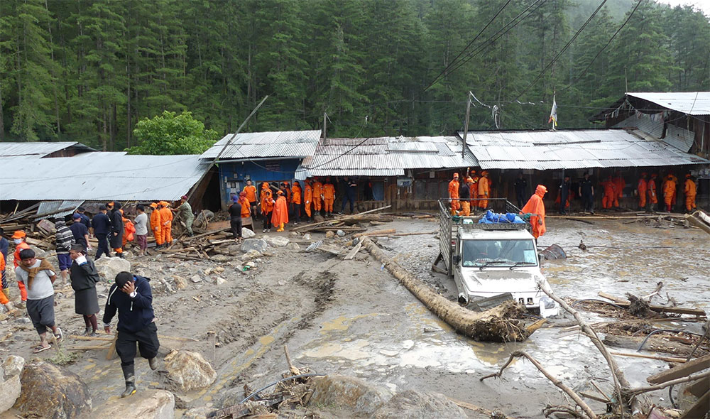 Photograph: muddy rural street by wooden sheds, orange-clad workers, and a small vehicle.